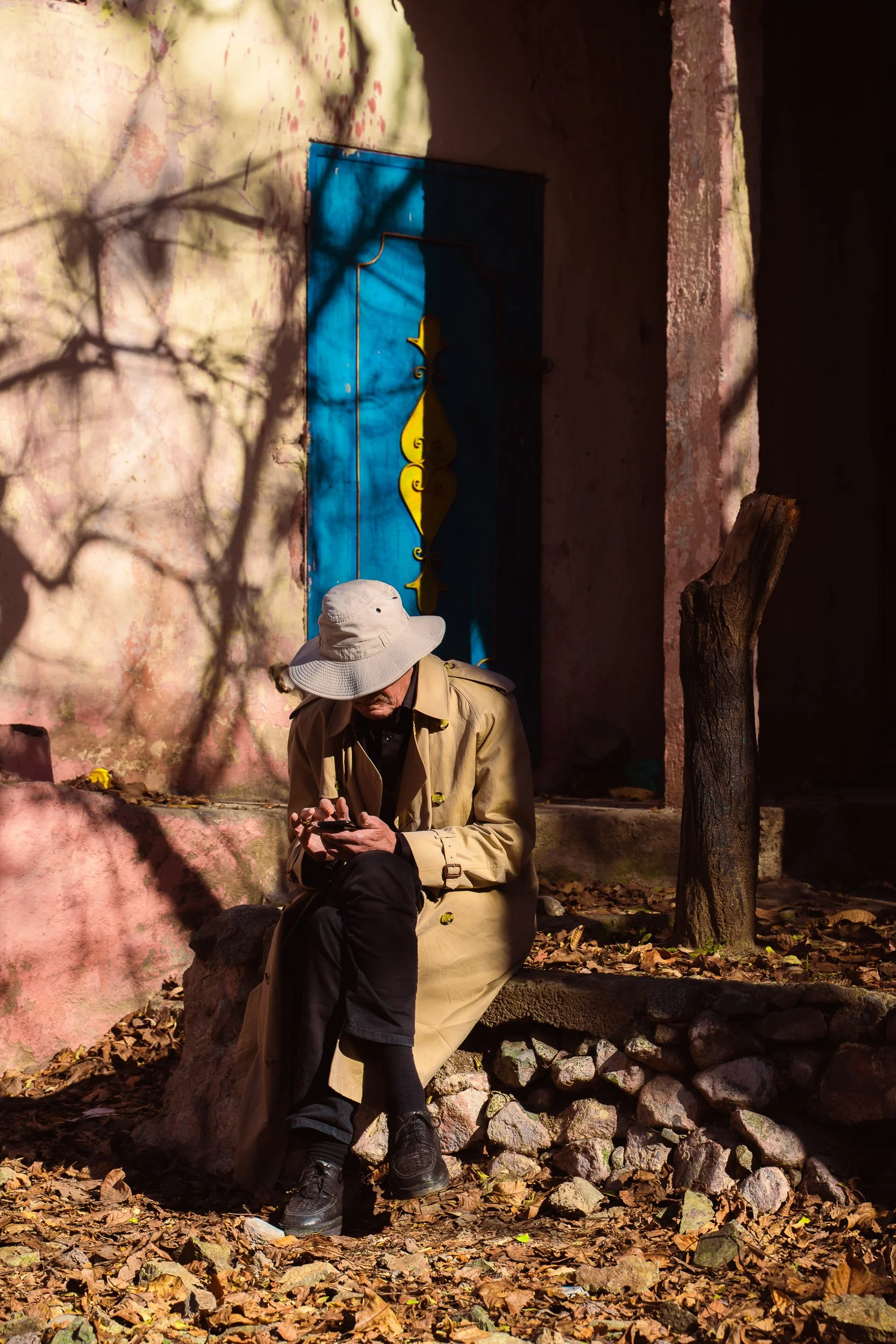 A man wearing a beige trench coat and a wide brimmed hat is sitting on a rock outdoors, looking at his phone. There are fallen leaves on the ground, a tree trunk, a textured pink wall, and a blue door behind him casting shadows.
