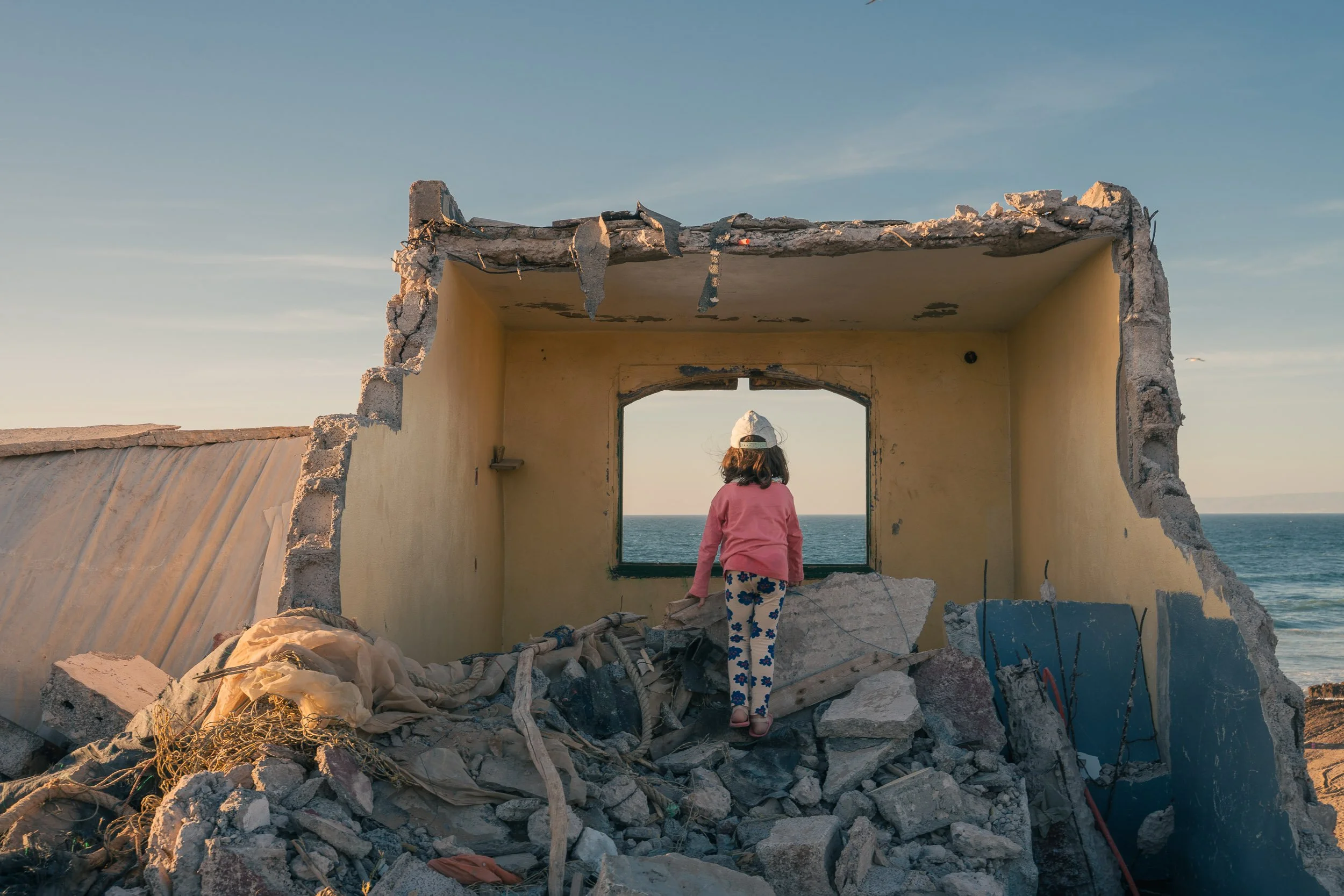 A young girl wearing a white cap, pink sweater, and patterned pants standing amidst the rubble of a destroyed building, looking out through a large open window at the ocean.
