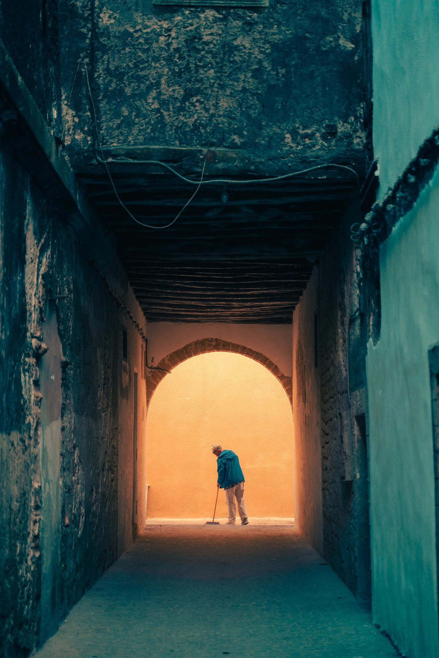 A person sweeping the floor in an arched alleyway with warm light at the end, surrounded by textured walls.