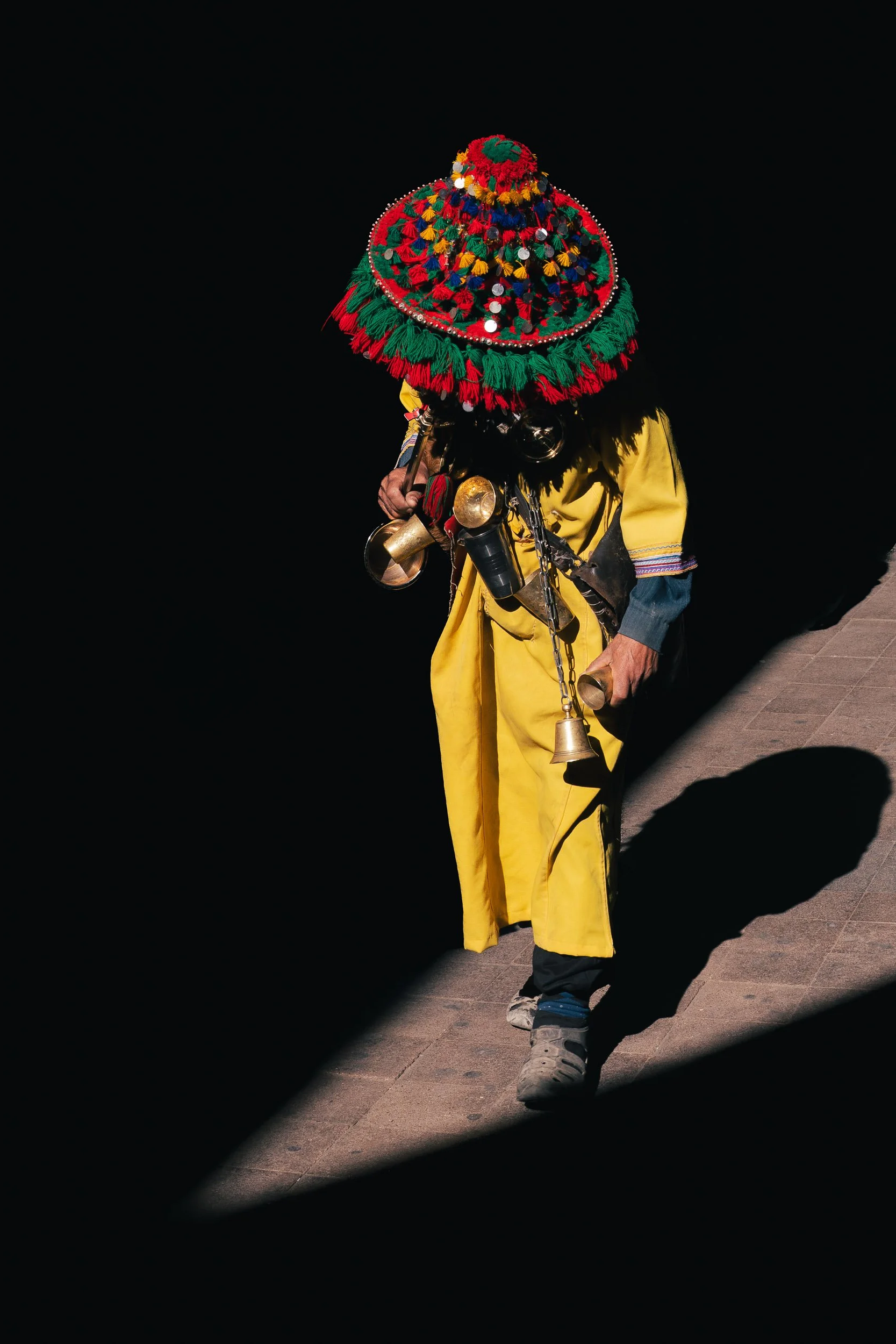 A person wearing a colorful, traditional Mexican hat and yellow clothing, holding small bells and pots, standing on a sidewalk at night with the shadow cast on the ground.