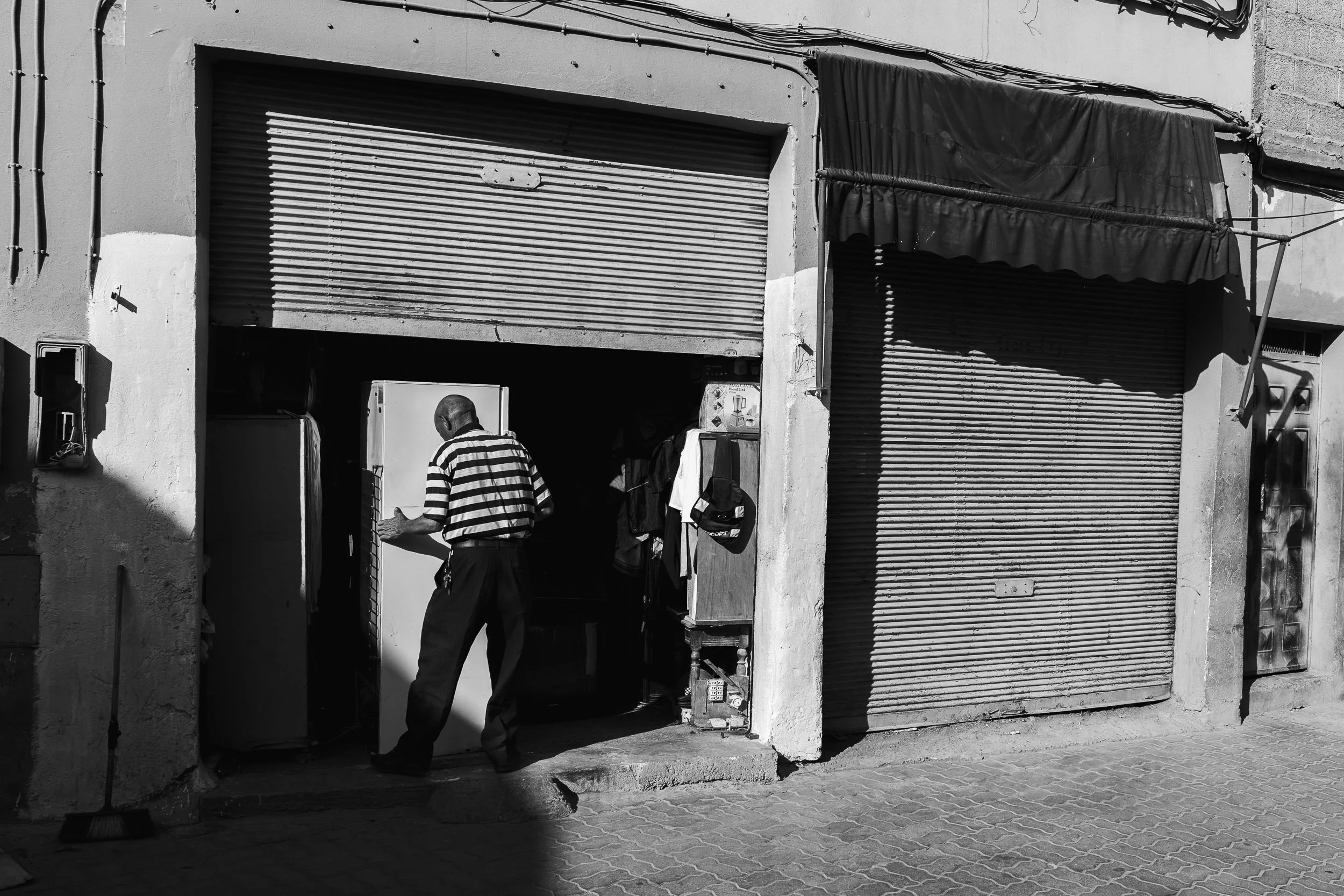 Man in striped shirt using an ATM outside a building with two metal roll-up doors and hanging clothing.