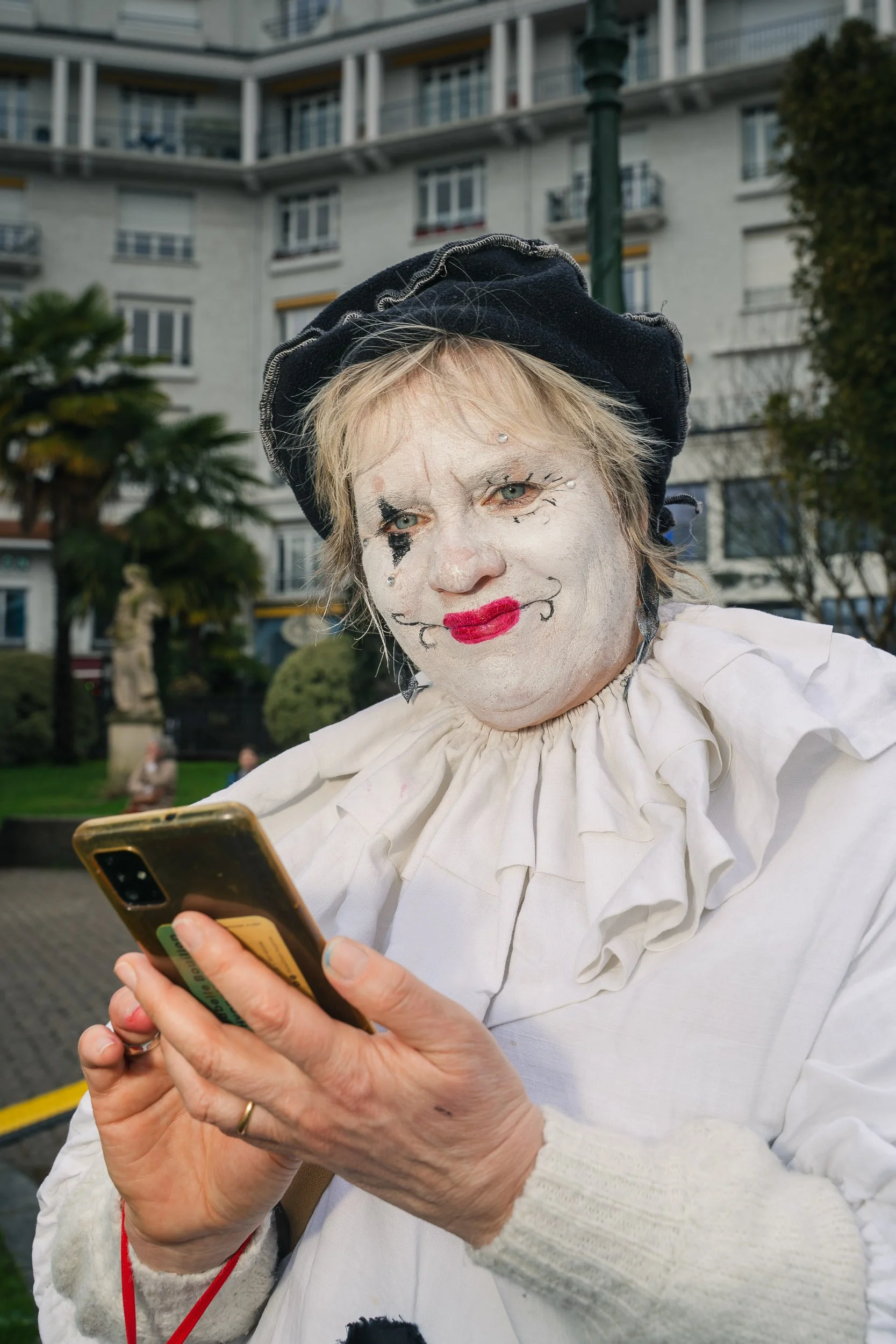 An elderly person with clown makeup on their face, wearing a black hat and a white ruffled blouse, looking at a smartphone in an outdoor urban setting.