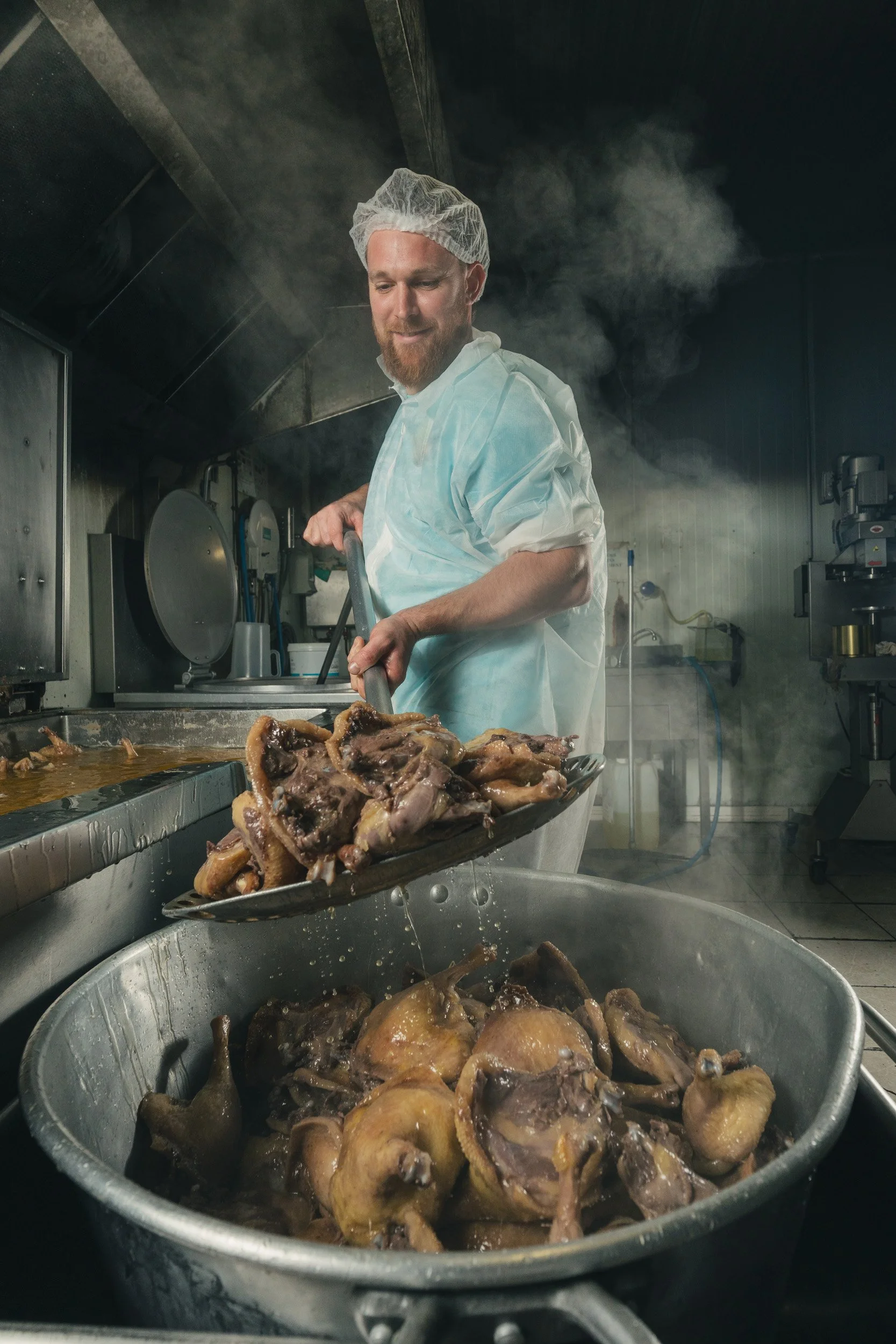 A chef wearing a white uniform and hairnet is preparing cooked poultry in a commercial kitchen, with steam rising in the background.