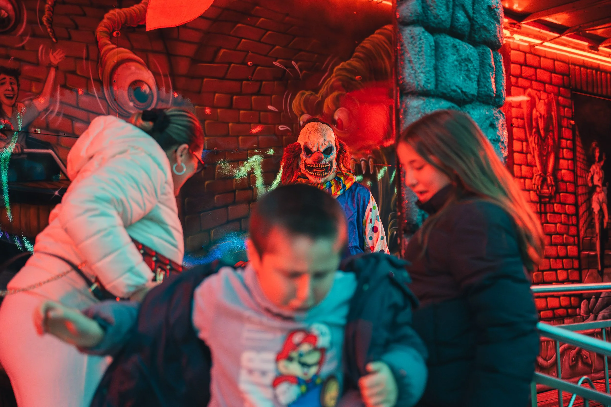 People on a spooky carnival ride with a clown face wall in the background, illuminated with red lights.