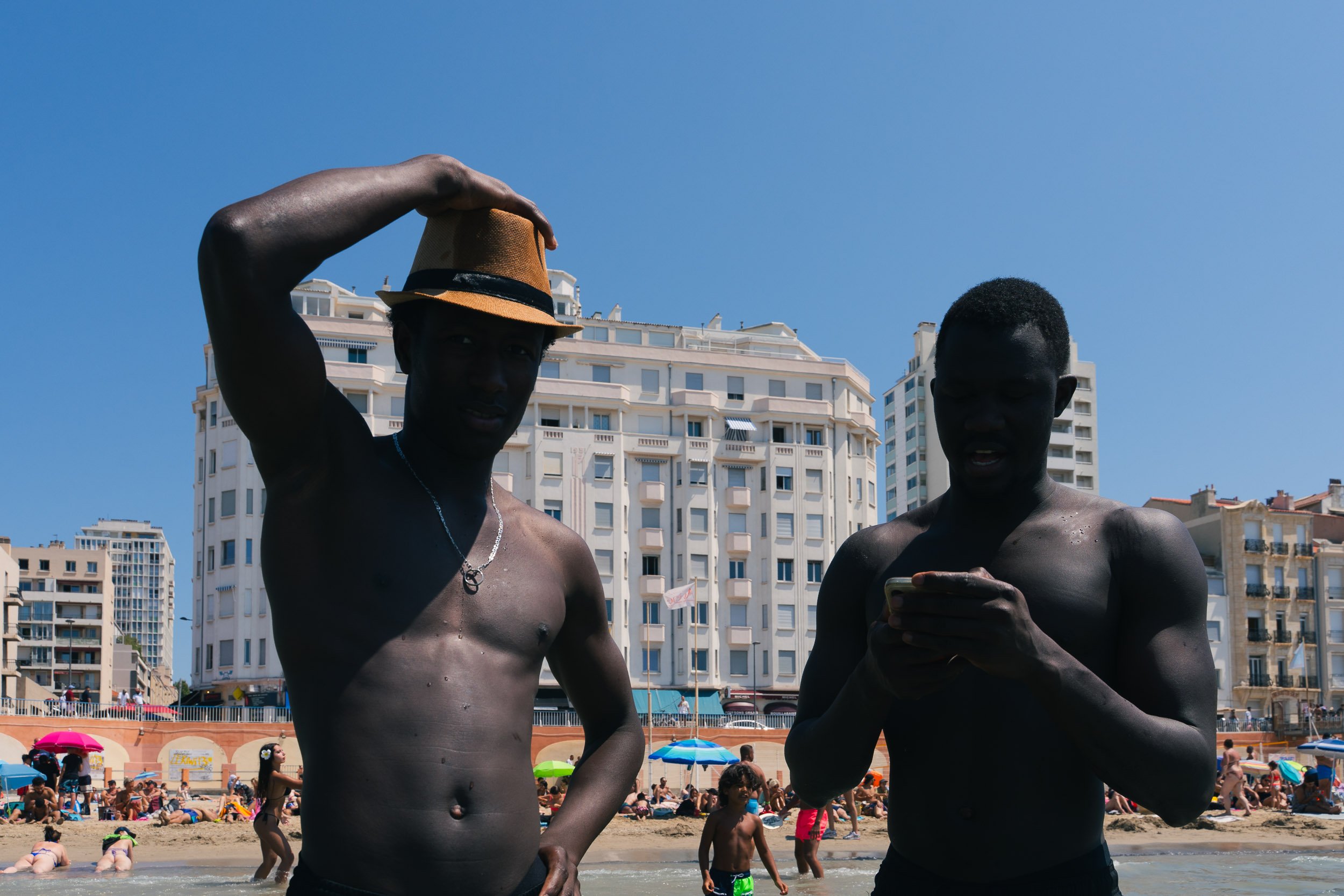 Two shirtless men standing in a crowded beach, one wearing a fedora hat and the other looking at a cellphone, with high-rise buildings and a blue sky in the background.