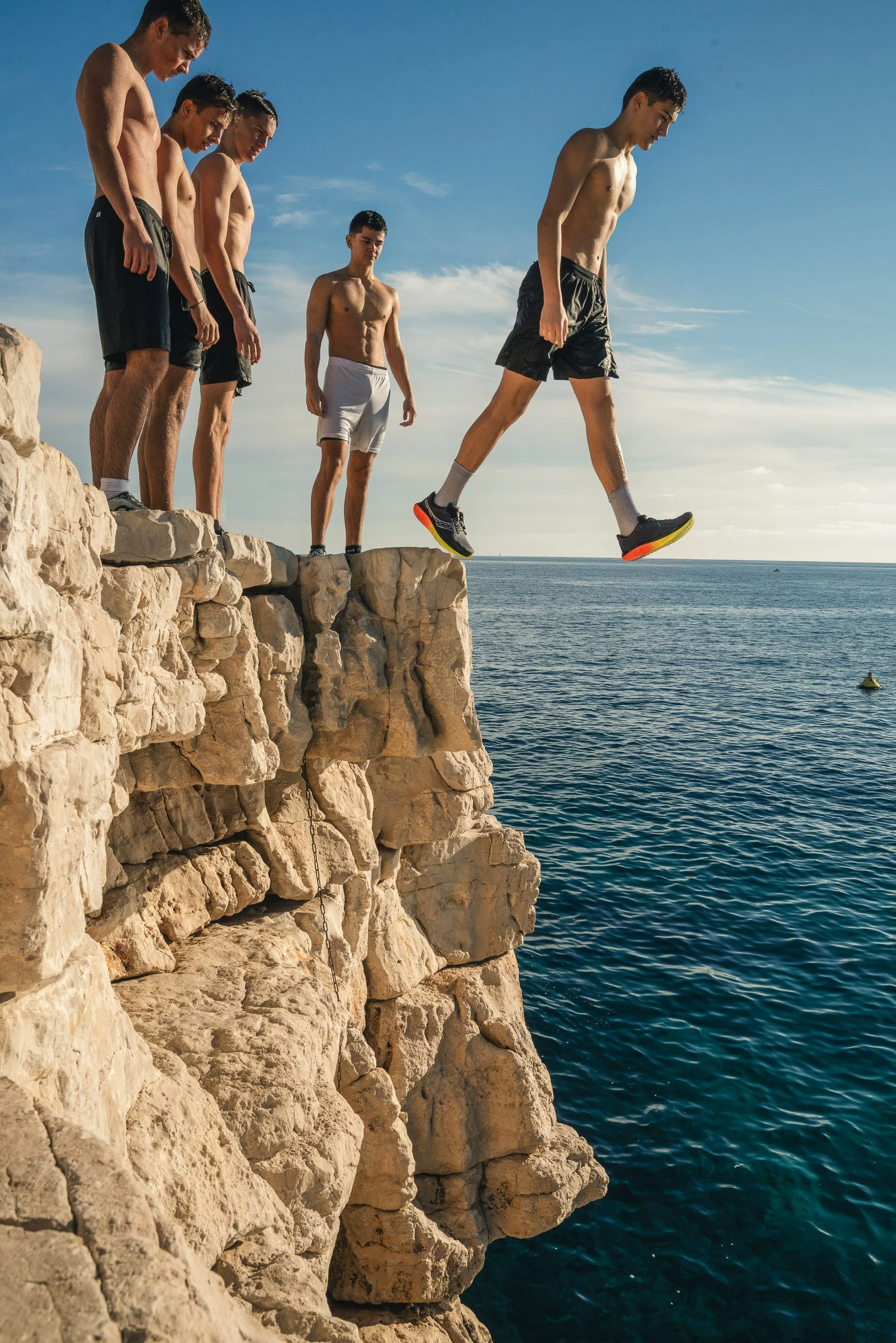 Five young men with athletic physiques in swim shorts standing on rocky cliff edge by the ocean, one is jumping off the edge into the water below during daylight.