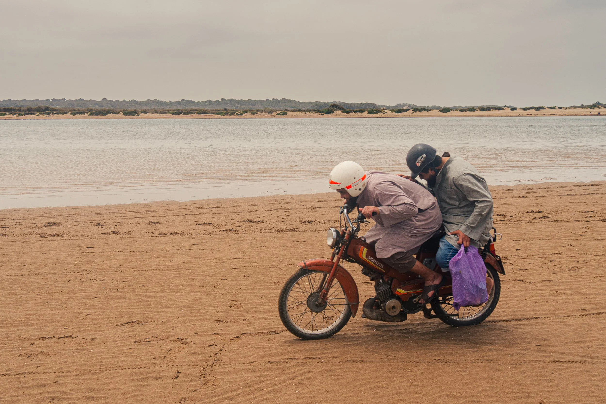 Two men riding a motorcycle with one in front and one seated behind, both wearing helmets, on a sandy beach next to a body of water, with a cloudy sky overhead.