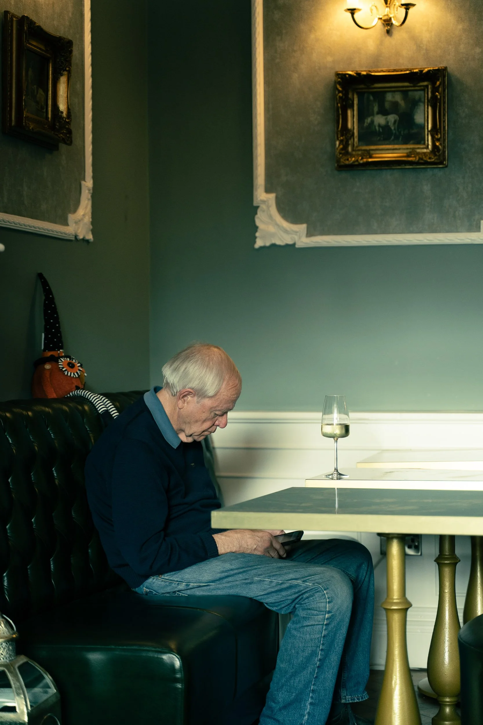 An elderly man with gray hair sits on a dark green leather bench, looking at his phone. A glass of white wine is on the table next to him, and framed paintings hang on the wall behind him.