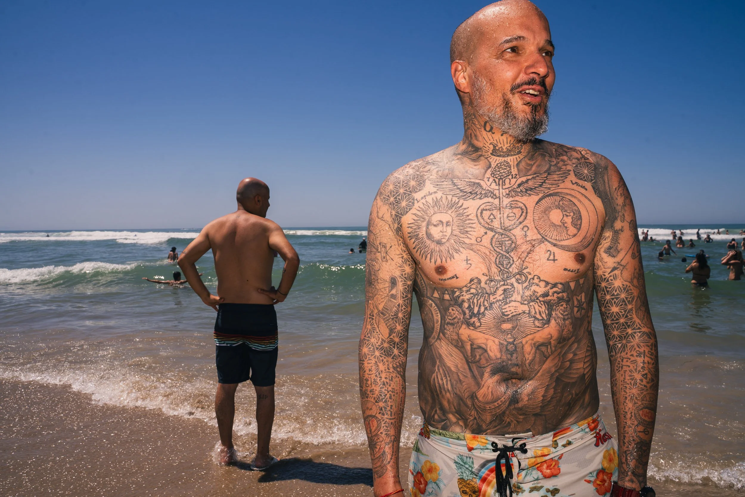 A middle-aged man with tattoos on his chest, arms, and neck stands on the beach, shirtless and smiling. In the background, another shirtless man with his hands on his hips looks at the ocean, where many people are swimming and playing in the waves un