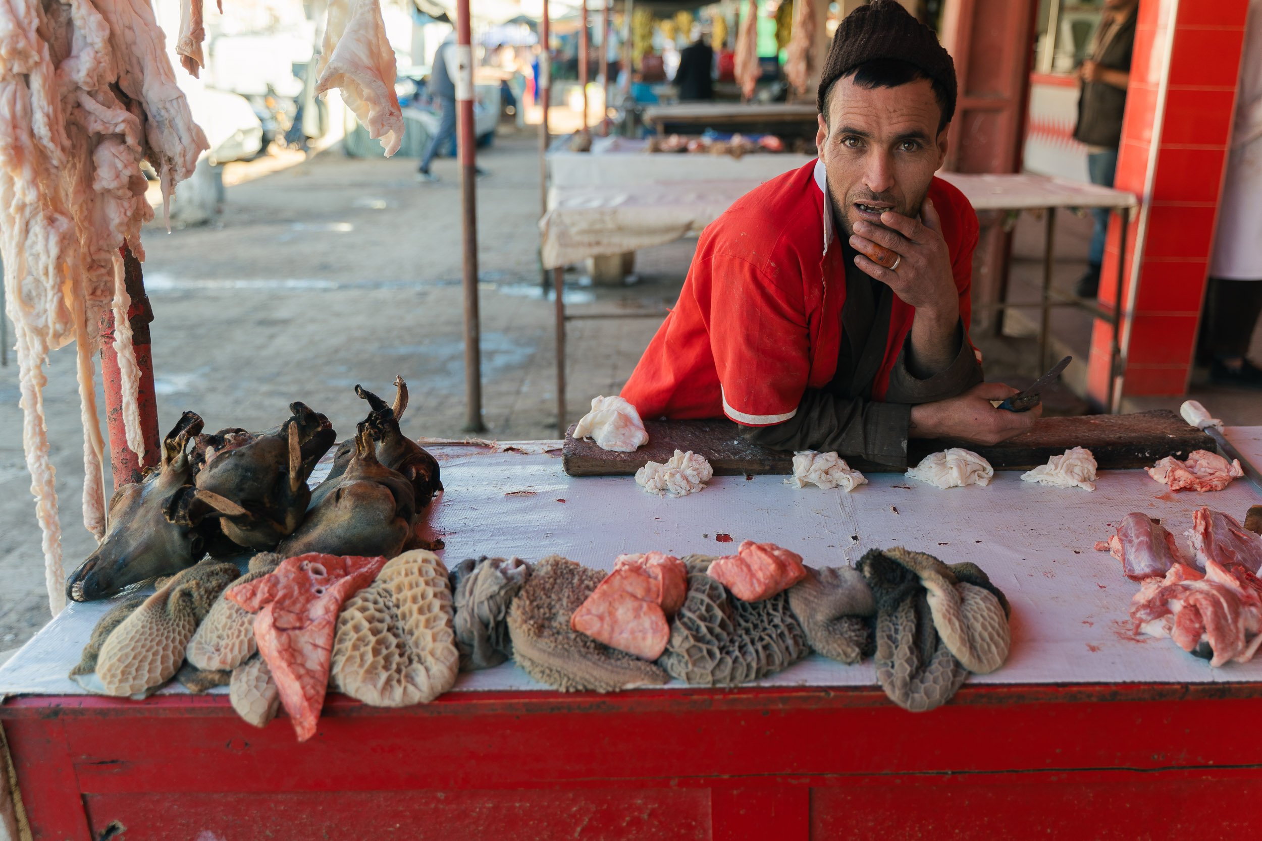 A man at a meat stall with animal heads, tripe, and cuts of meat on the counter, outdoors on a market street.