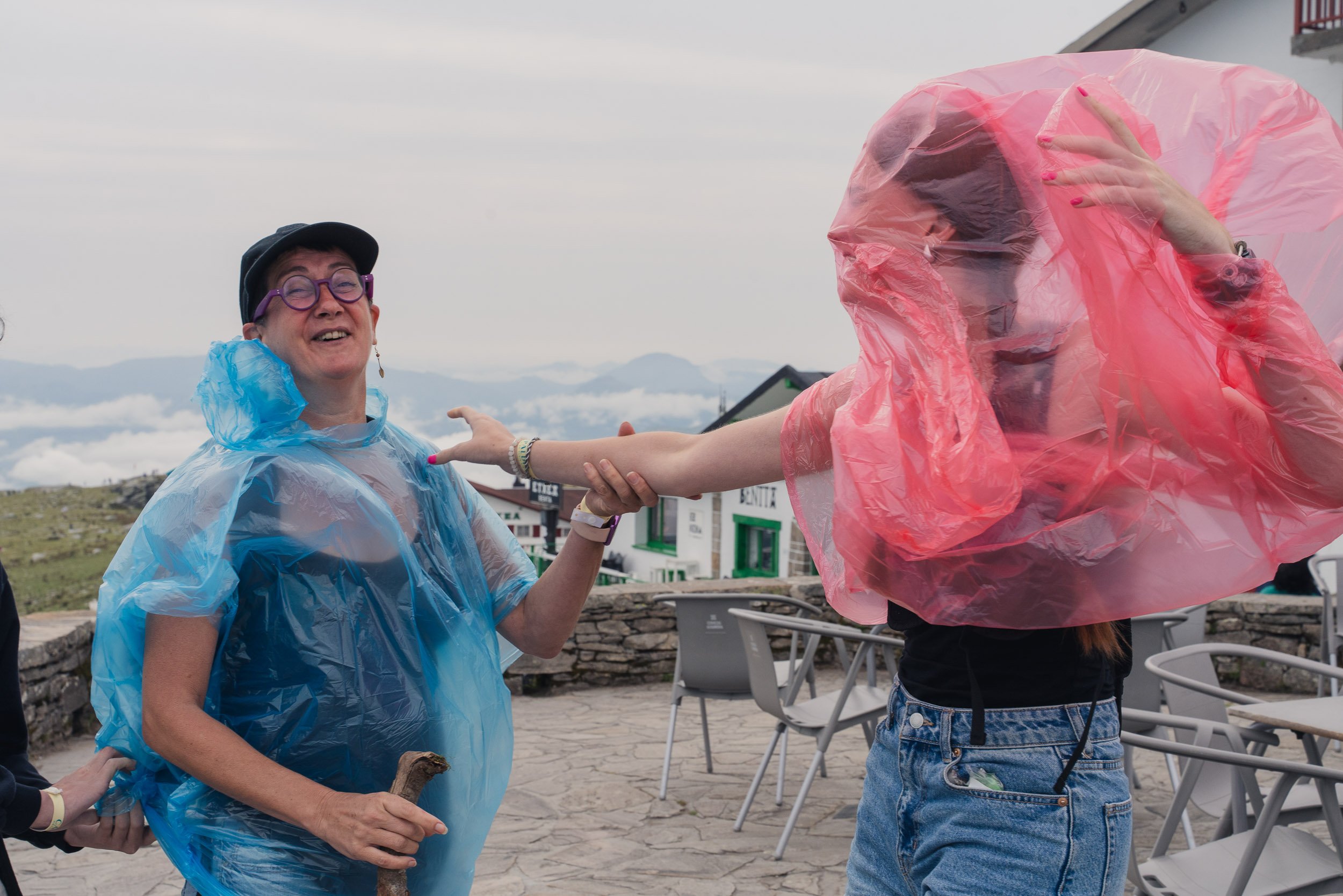 Two women wearing pink and blue rain ponchos are outside with mountains and buildings in the background. One woman is smiling and holding a walking stick, while the other is reaching out her hand. The sky is cloudy.