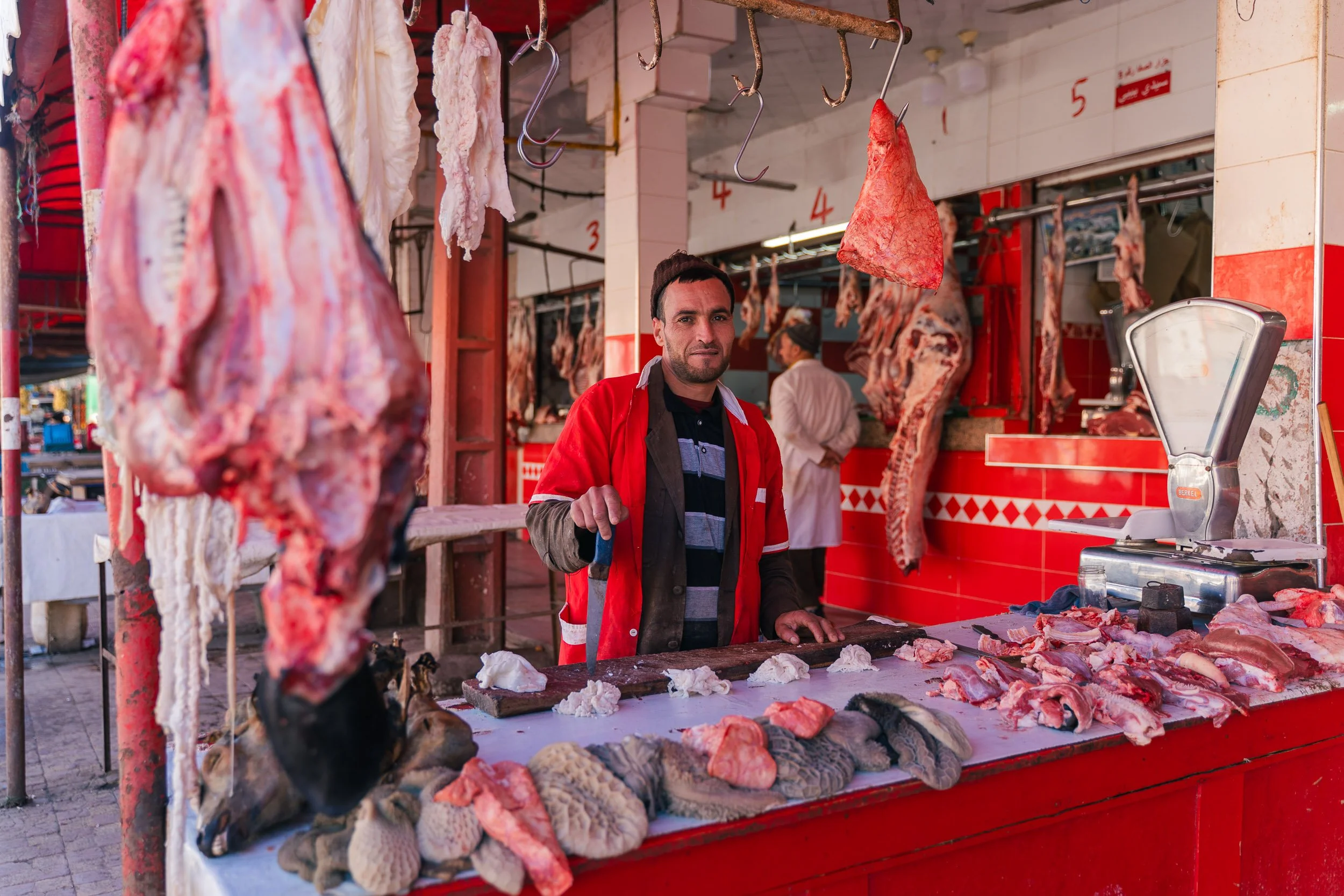 Market butcher stall with various cuts of meat displayed on a counter, hanging meats above, and a man in a red coat behind the counter.