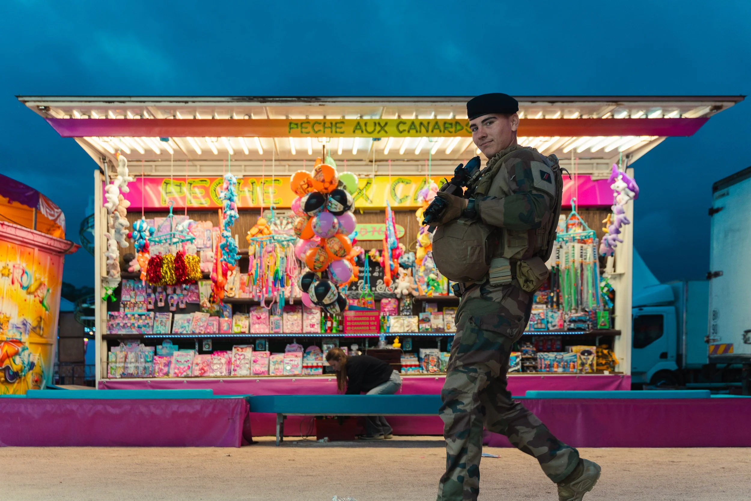 A young man in military camouflage uniform holding a camera walking past a colorful carnival game booth labeled "FEECHE AUX CANARD" with stuffed duck toys and hanging prizes inside.