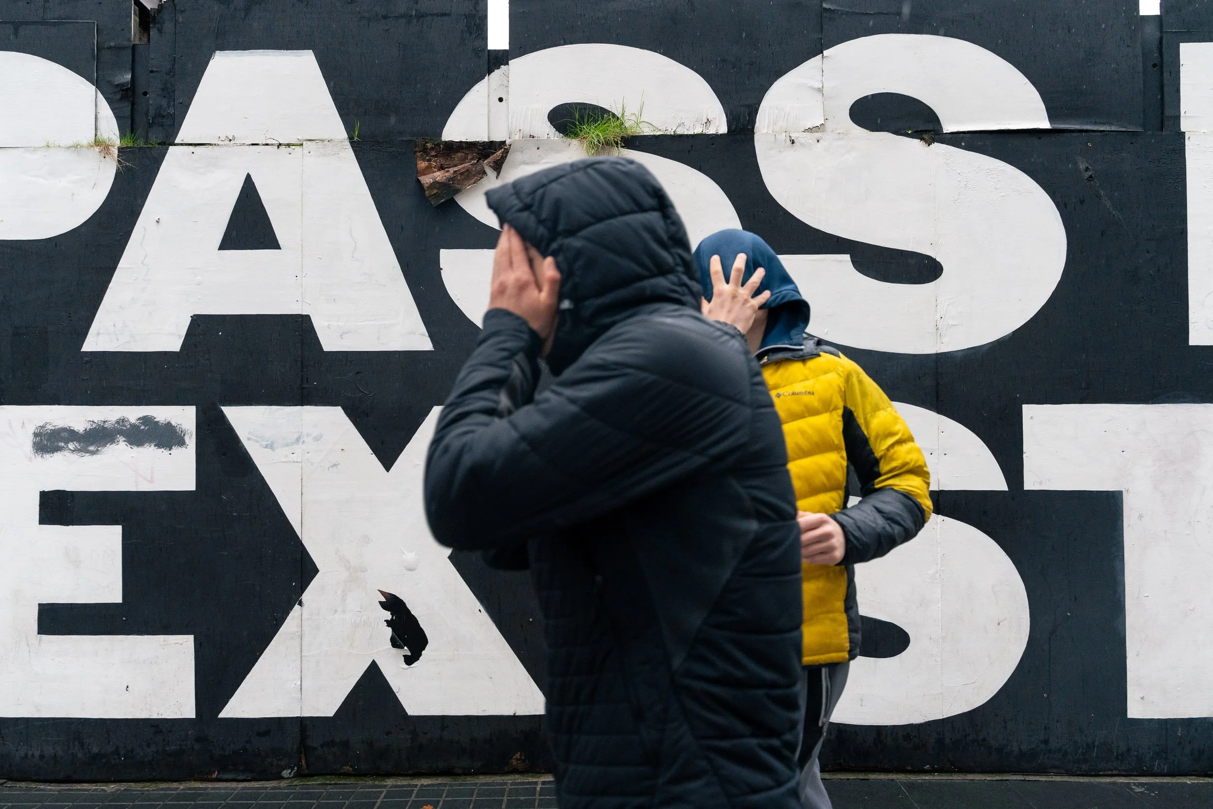 Two people walking past a large black and white wall with bold letters. One person is in a black jacket with the hood up, covering their face with their hands. The other person is in a yellow and gray jacket, facing downward.