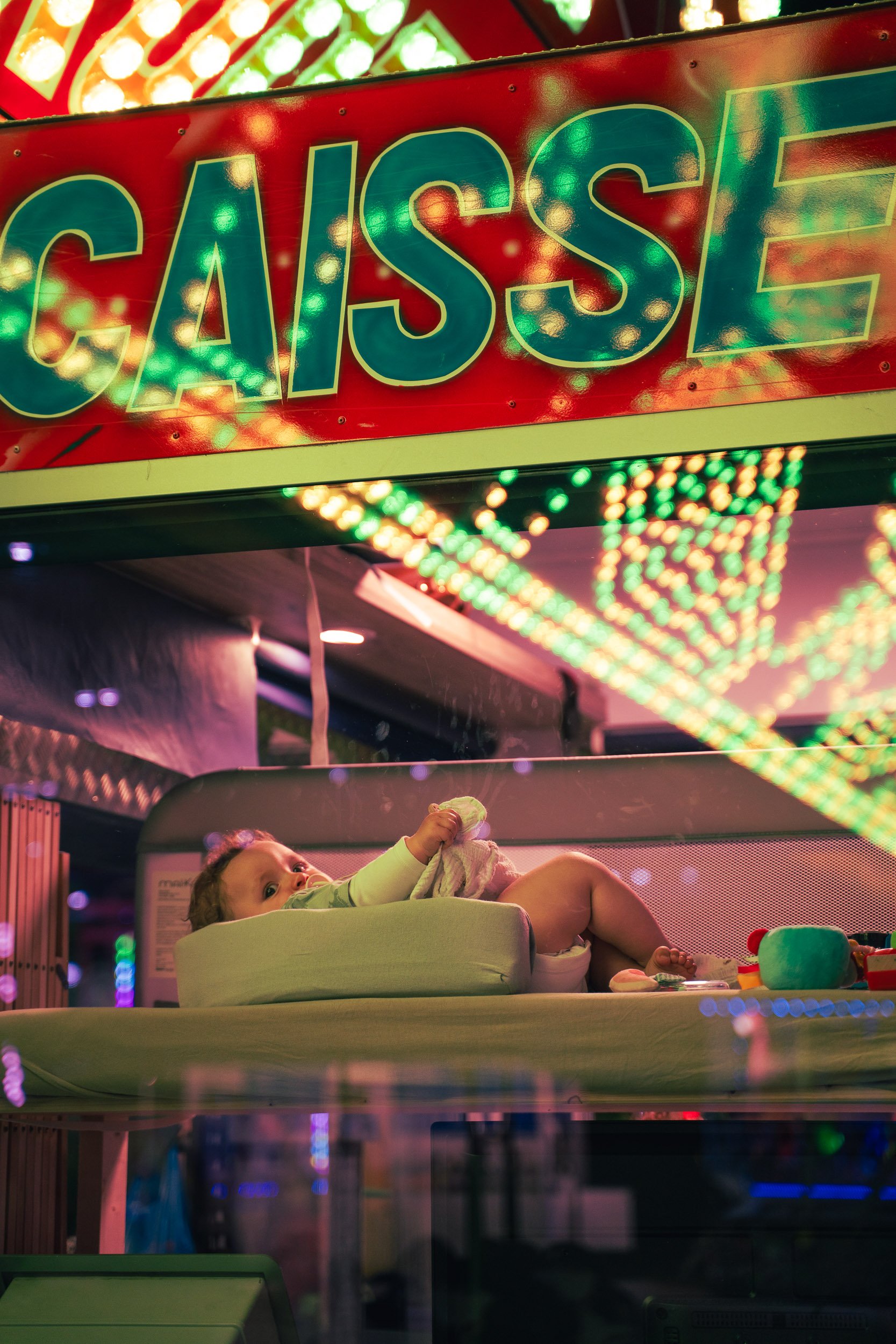 A young child lying on a bed inside a carnival game's prize area, with bright, colorful neon lights and a large sign that says 'CASS' behind them.