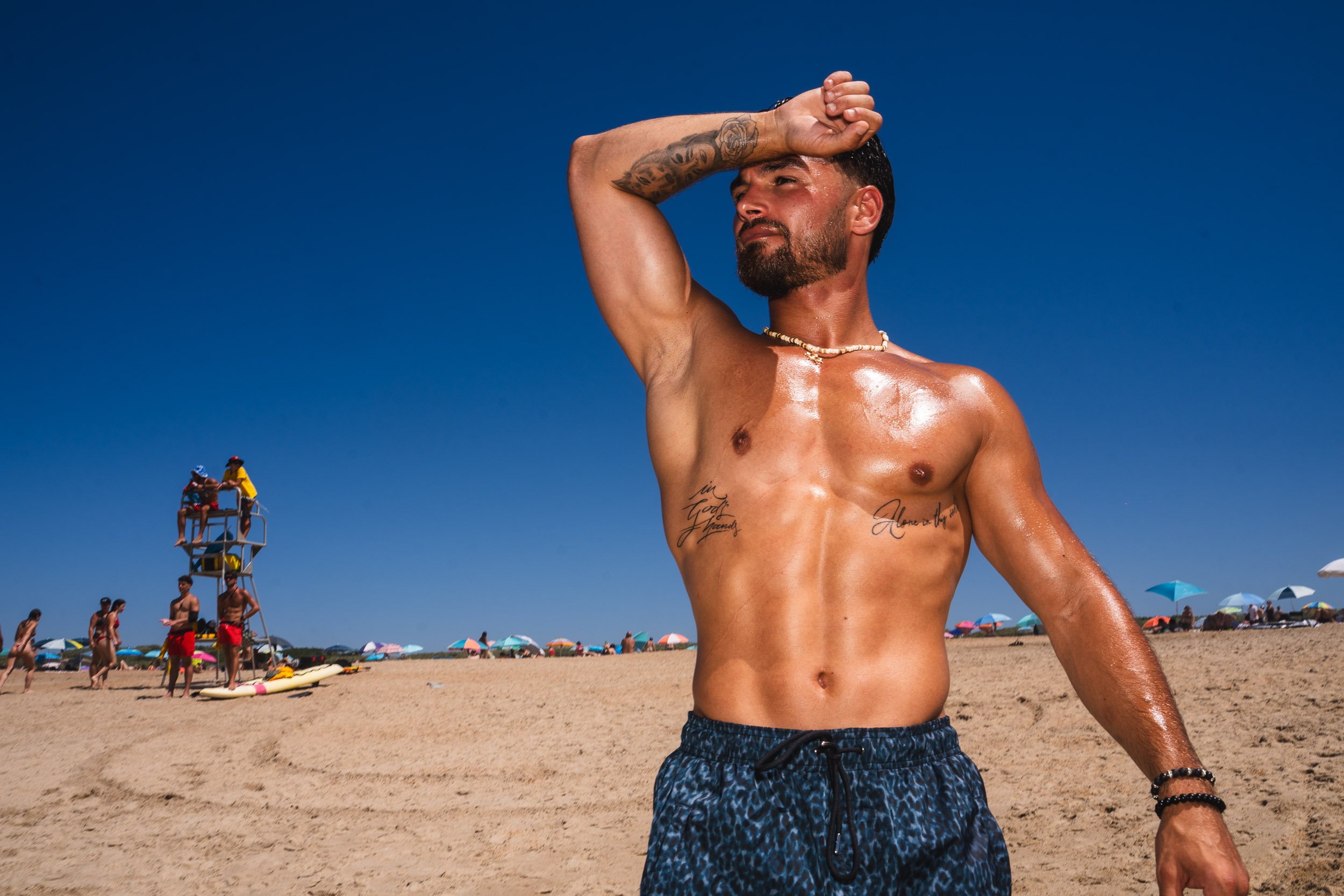 A muscular man with tattoos on his chest and arm, wearing swimming shorts and a shell necklace, shielding his eyes from the sun on a beach with umbrellas and people in the background.