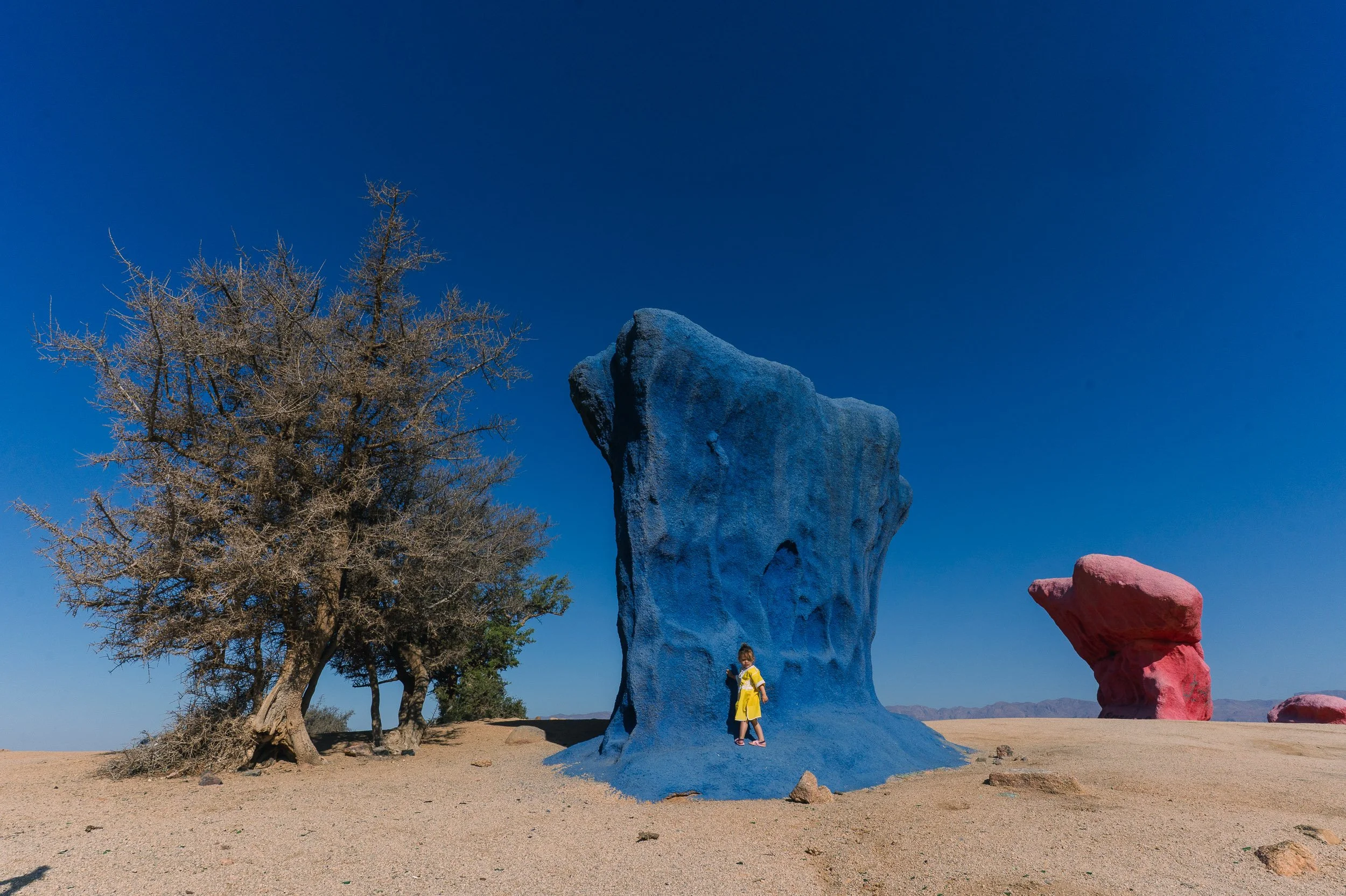 A girl in a yellow dress standing next to a large blue rock formation with a child-sized figure on top, in a desert landscape with leafless trees, large red rocks, and a clear blue sky.