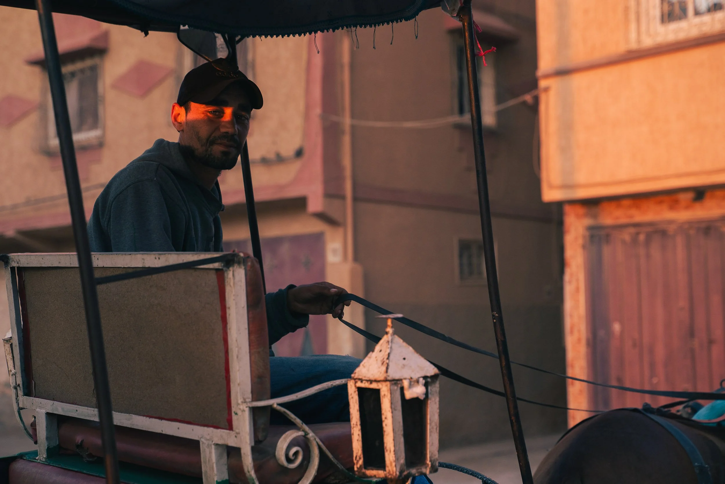 A man sitting in a horse-drawn carriage during sunset, wearing a black cap and hoodie, with a background of pink and orange buildings.