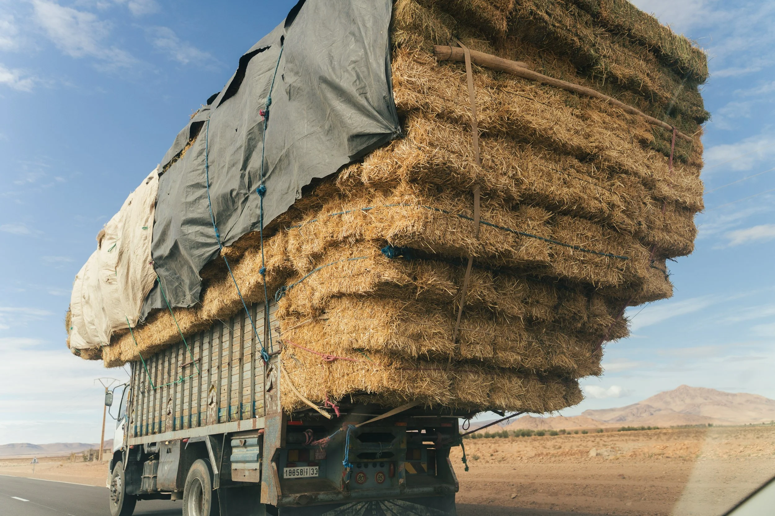 A truck on a highway carrying a large load of hay bales stacked high in a desert landscape with mountains in the background.