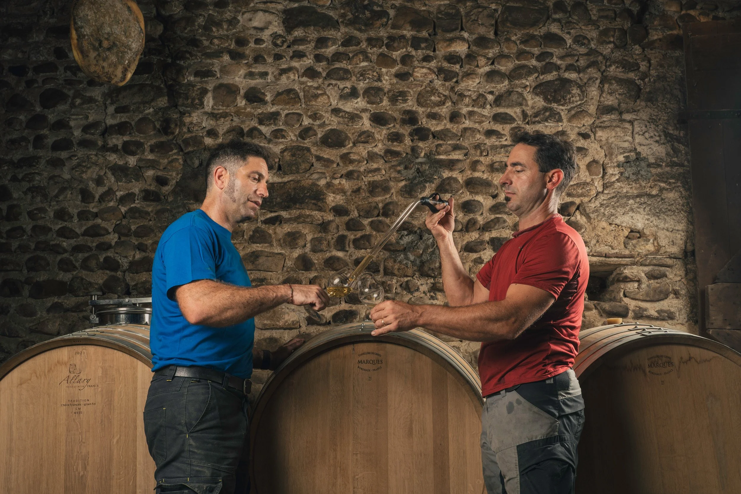 Two men pouring wine in a cellar with stone walls and large wooden wine barrels.