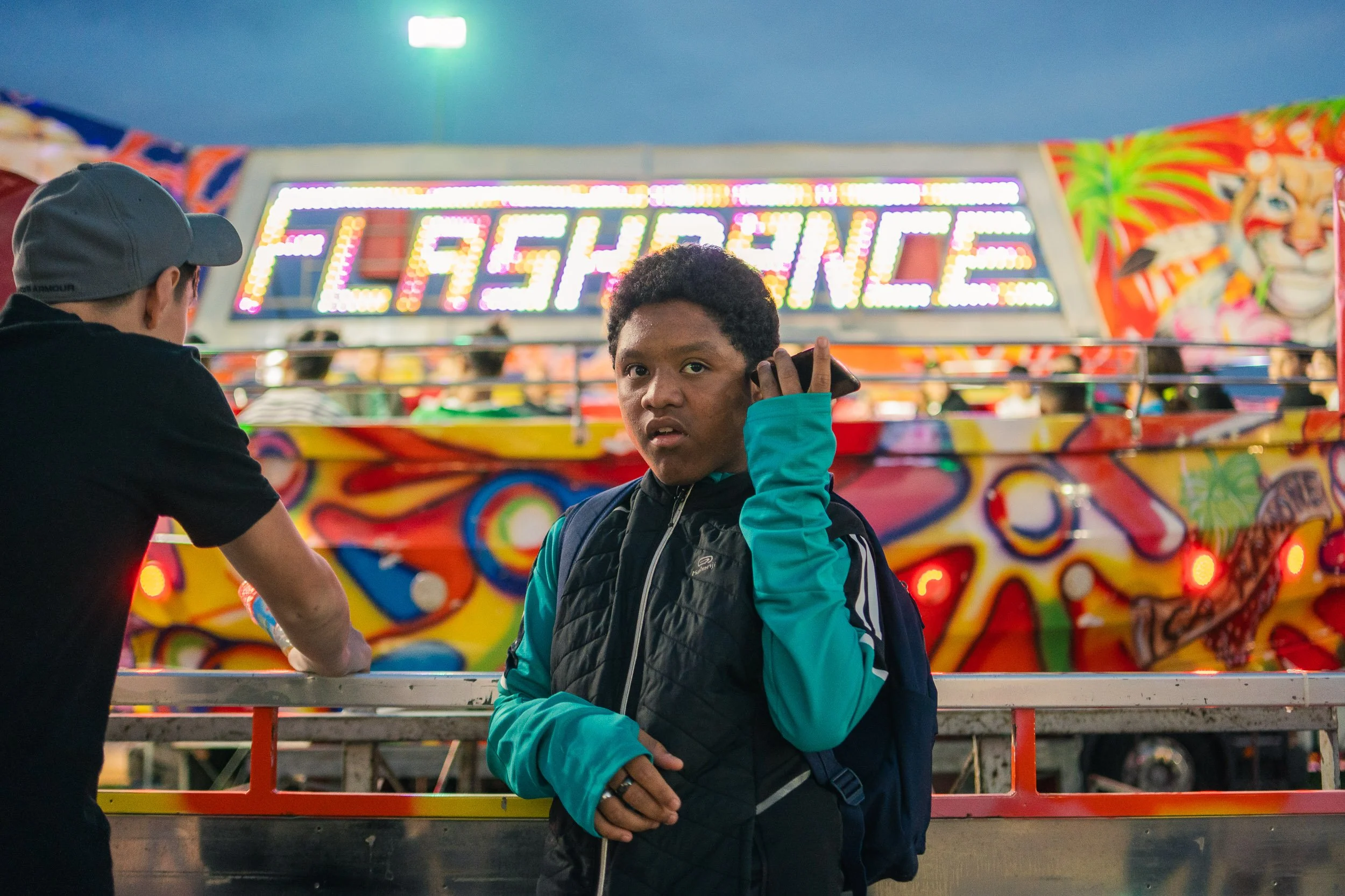 A young man in a teal jacket and black vest holding a phone to his ear, standing in front of a brightly lit carnival ride with a sign that reads "LASSENGE."