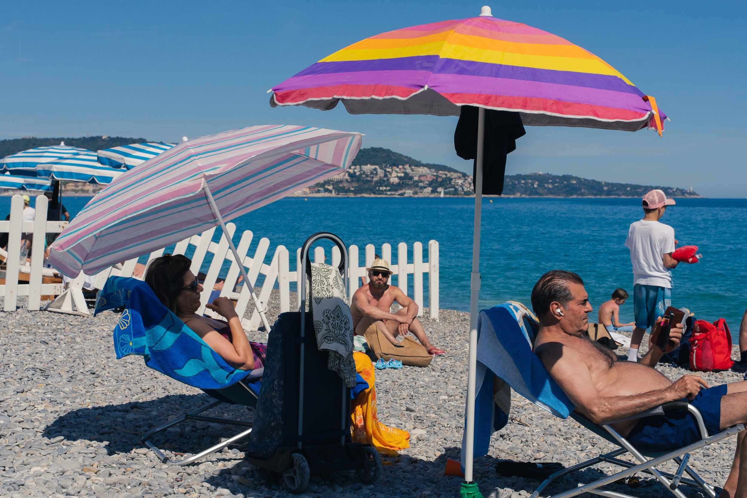 People relaxing on a pebble beach under colorful umbrellas, with the sea and hills in the background.