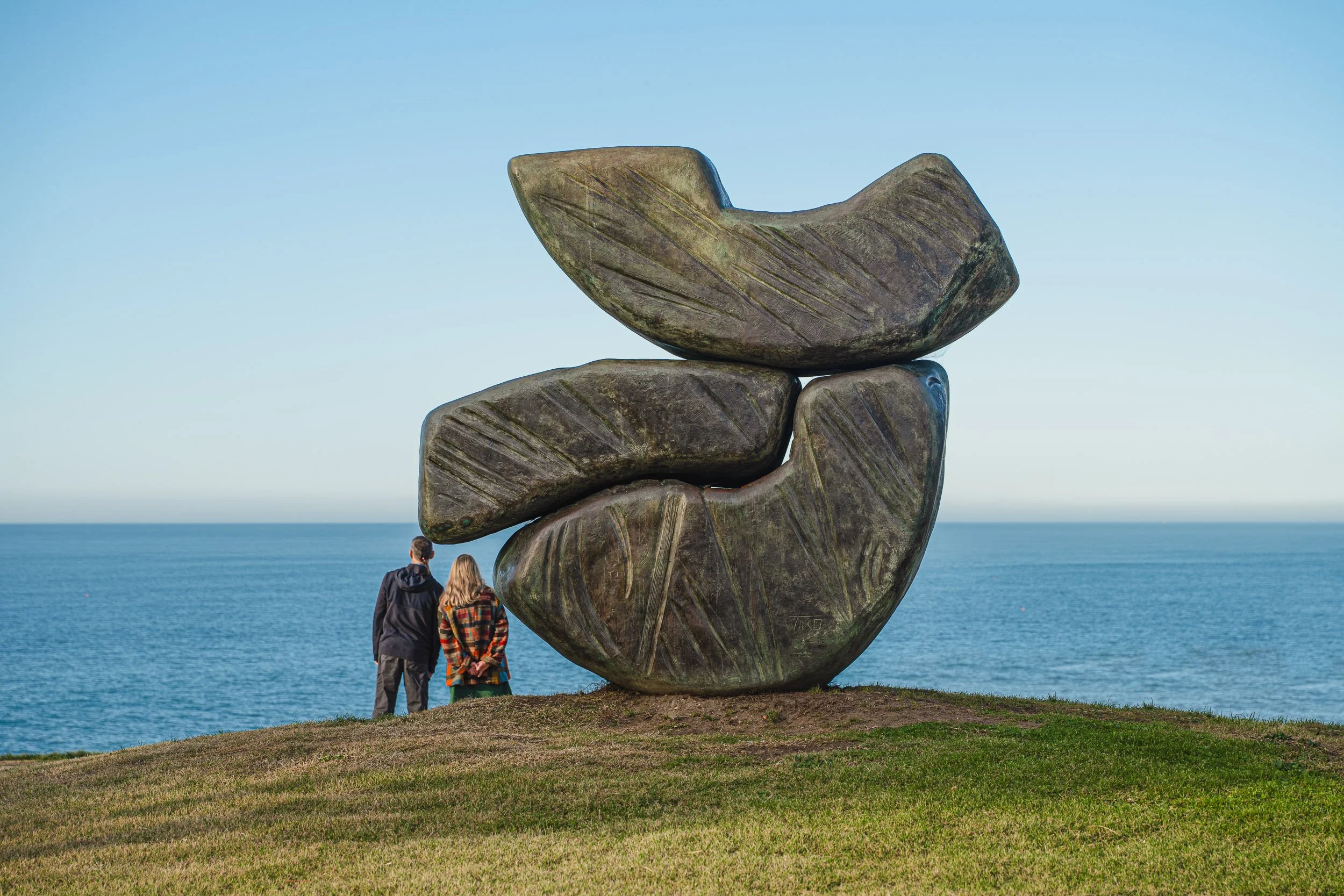 Two people, a man and a woman, standing on a grassy area near the sea, looking at a large abstract stone sculpture composed of three large stacked rocks.