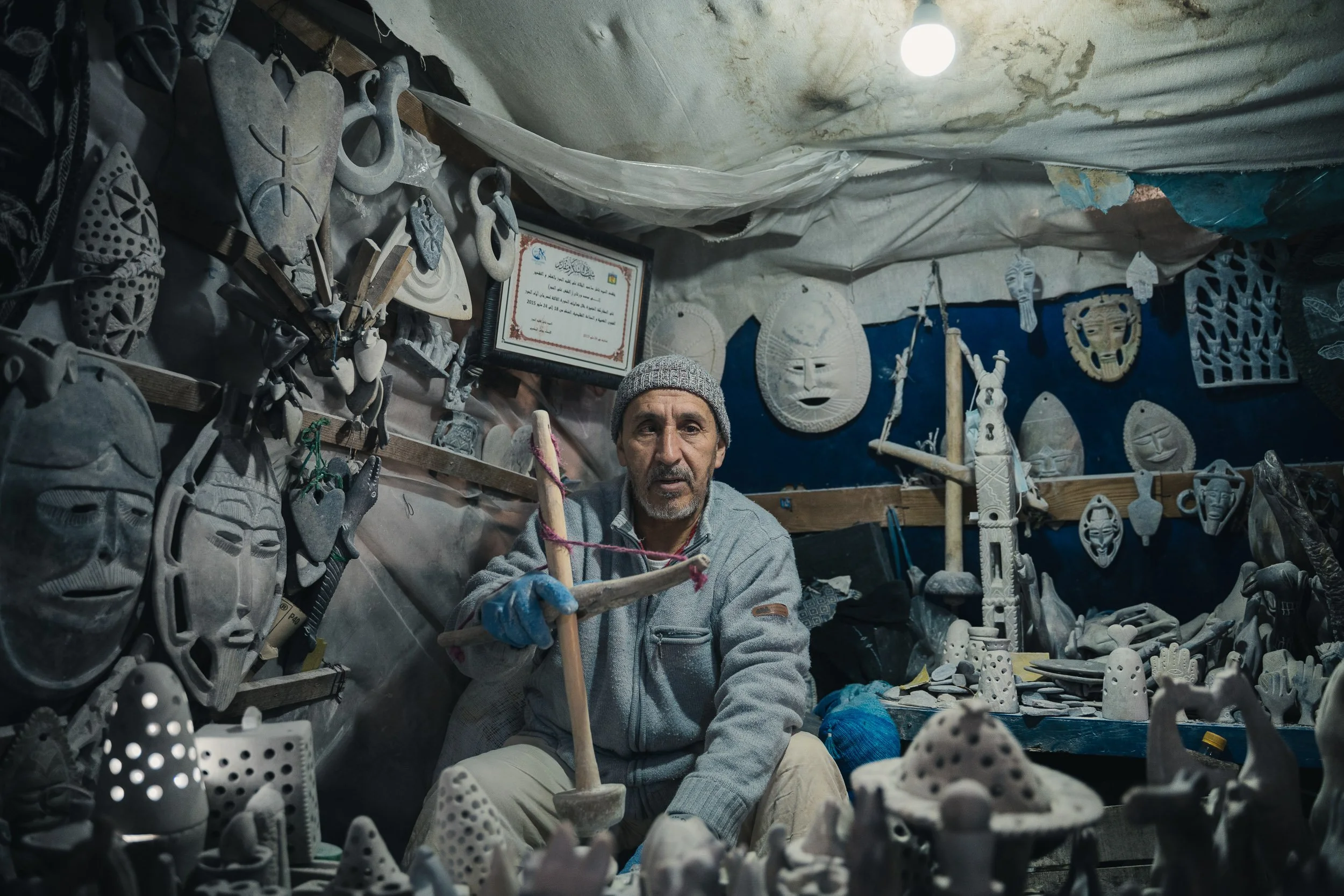 A man wearing a gray beanie and gray jacket sits in a workshop filled with traditional carved masks and sculptures. The workshop has fabric covering the walls and ceiling, with various masks and carved items hanging and displayed around him.