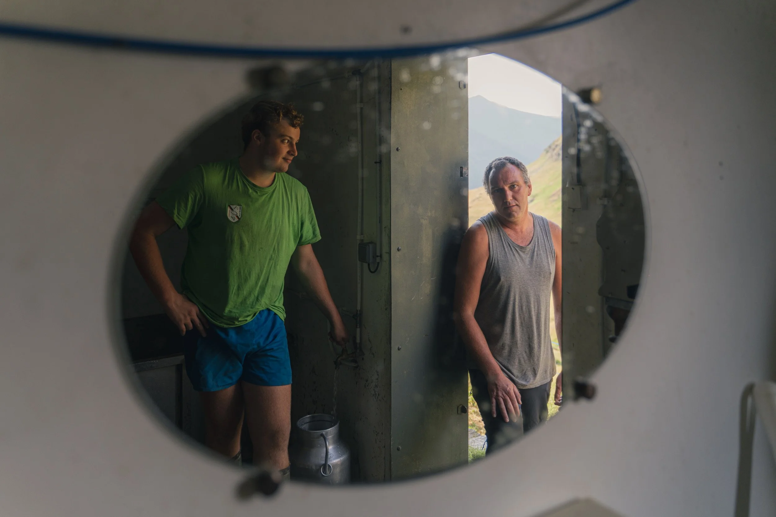 Two men standing outside a building seen through a circular window, with one wearing a green shirt and blue shorts, and the other in a gray tank top, in a rural landscape with mountains in the background.