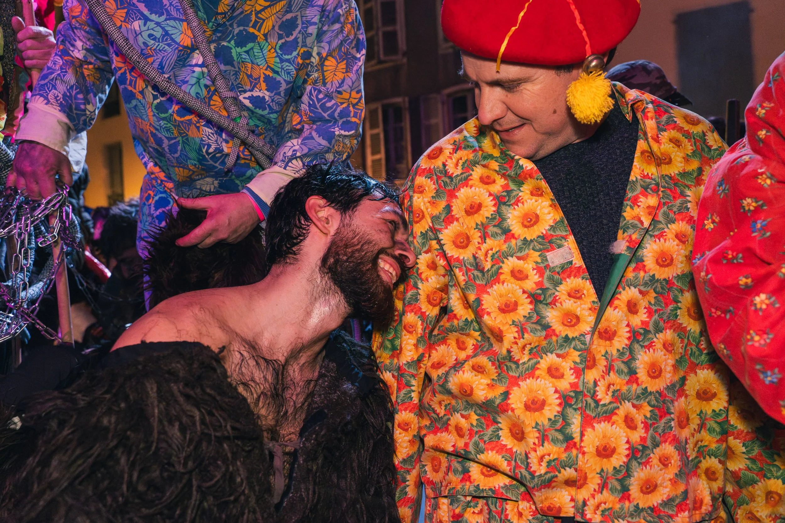 People dressed in colorful costumes at a festive event, with one person appearing emotional or joyful, lying backward with their head tilted and facial expression showing happiness or relief, surrounded by others in celebratory attire.