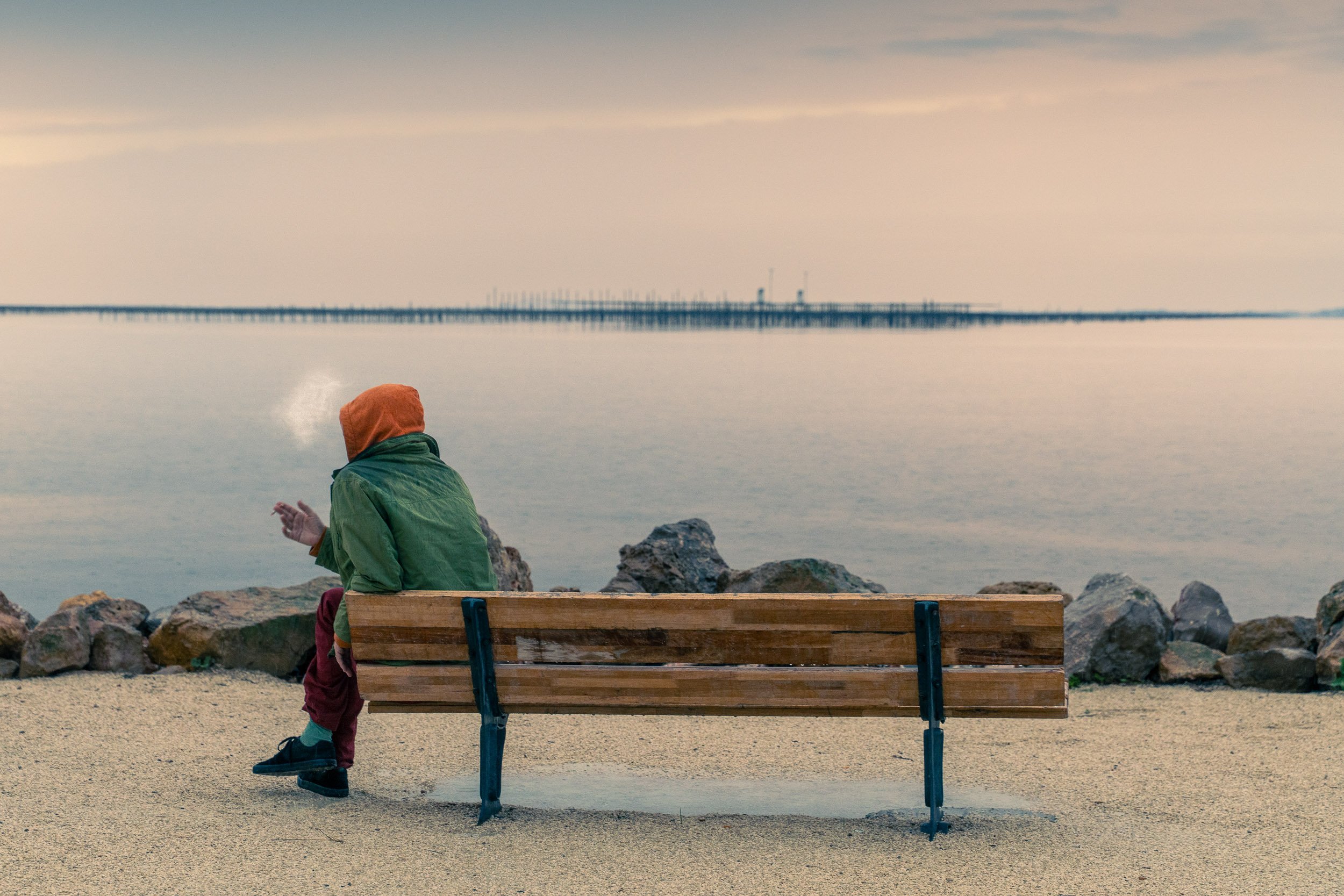 A person sitting on a wooden bench near the water, wearing an orange hooded jacket and green coat, with rocks and a calm body of water in the background.