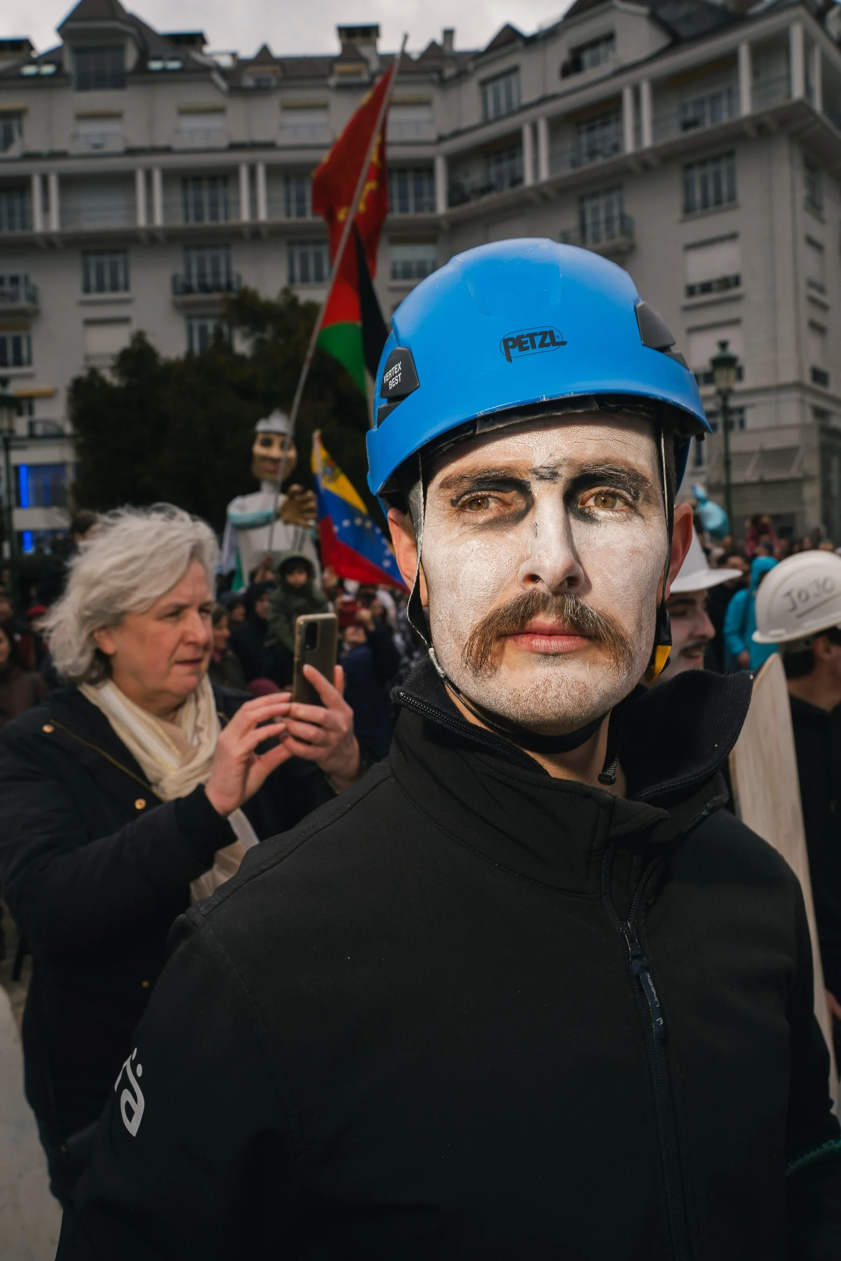 A man wearing a blue Petzl helmet with face paint resembling a skeleton, participating in a protest or demonstration, with a crowd and flags in the background.