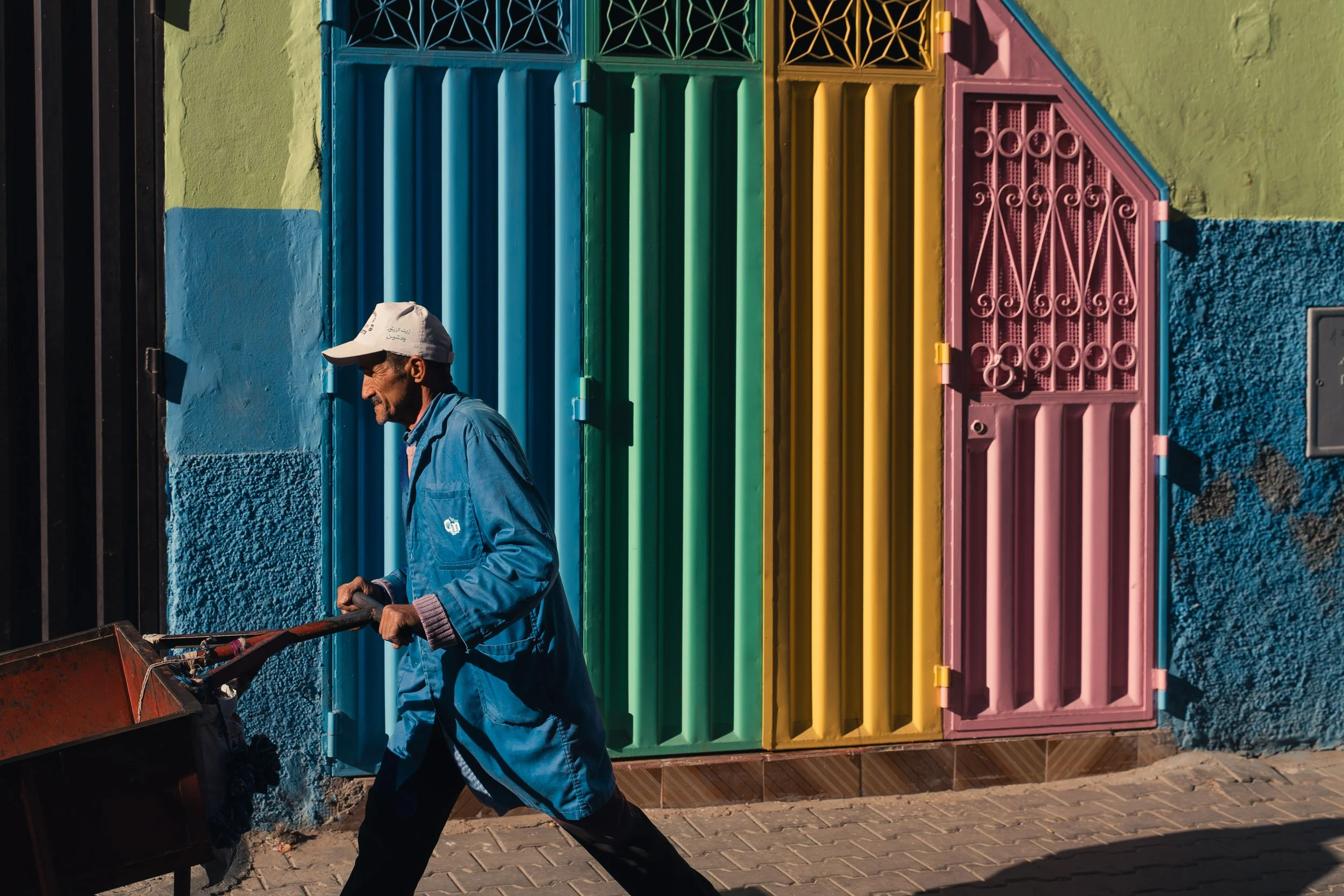 Un homme portant une casquette blanche et une veste bleue pousse une brouette, devant un portail coloré composé de sections peintes en bleu, vert, jaune et rose, à Taroudant - Maroc
