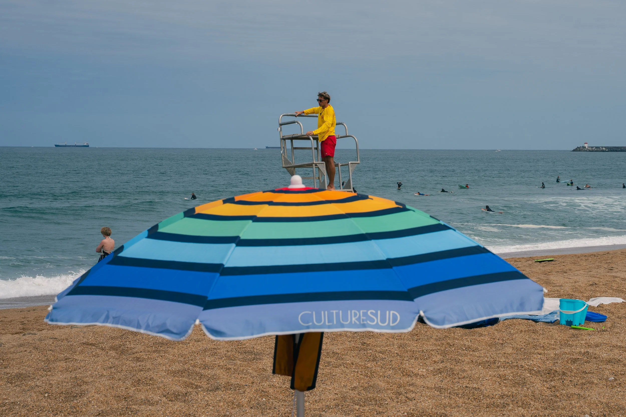 Beach scene with colorful umbrella in the foreground, swimmers in the water, a lifeguard on the tower, and a cloudy sky.