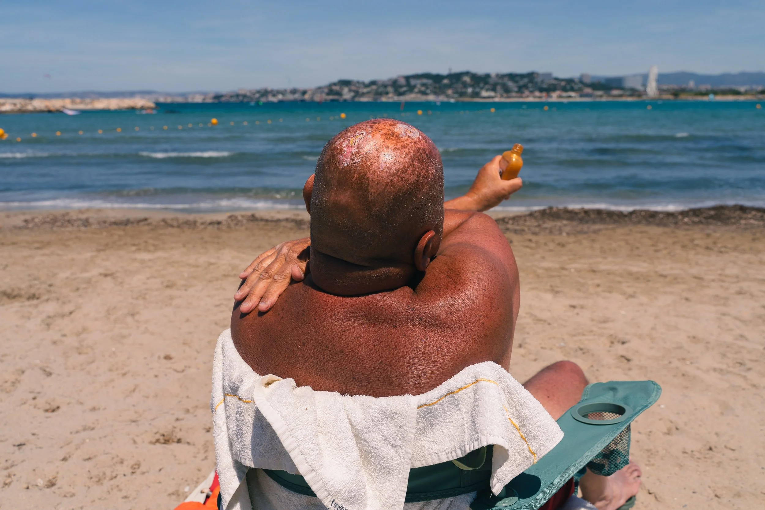 An elderly man with a bald head sits on a beach chair, facing the ocean, holding a small bottle of sunscreen.
