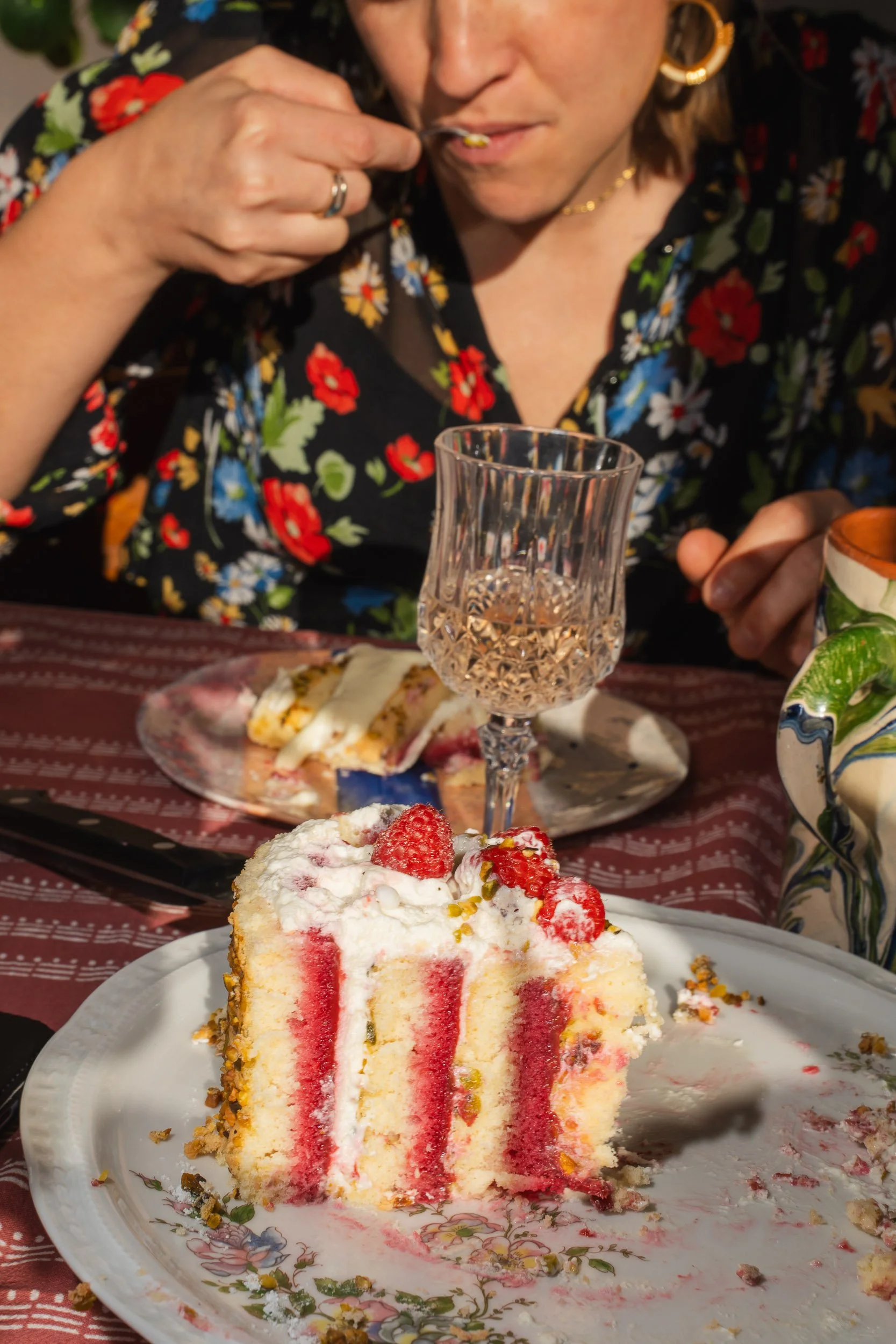 A woman in a black floral dress eating a slice of cake with strawberries, on a decorated party table with a crystal glass, plates, and other desserts.
