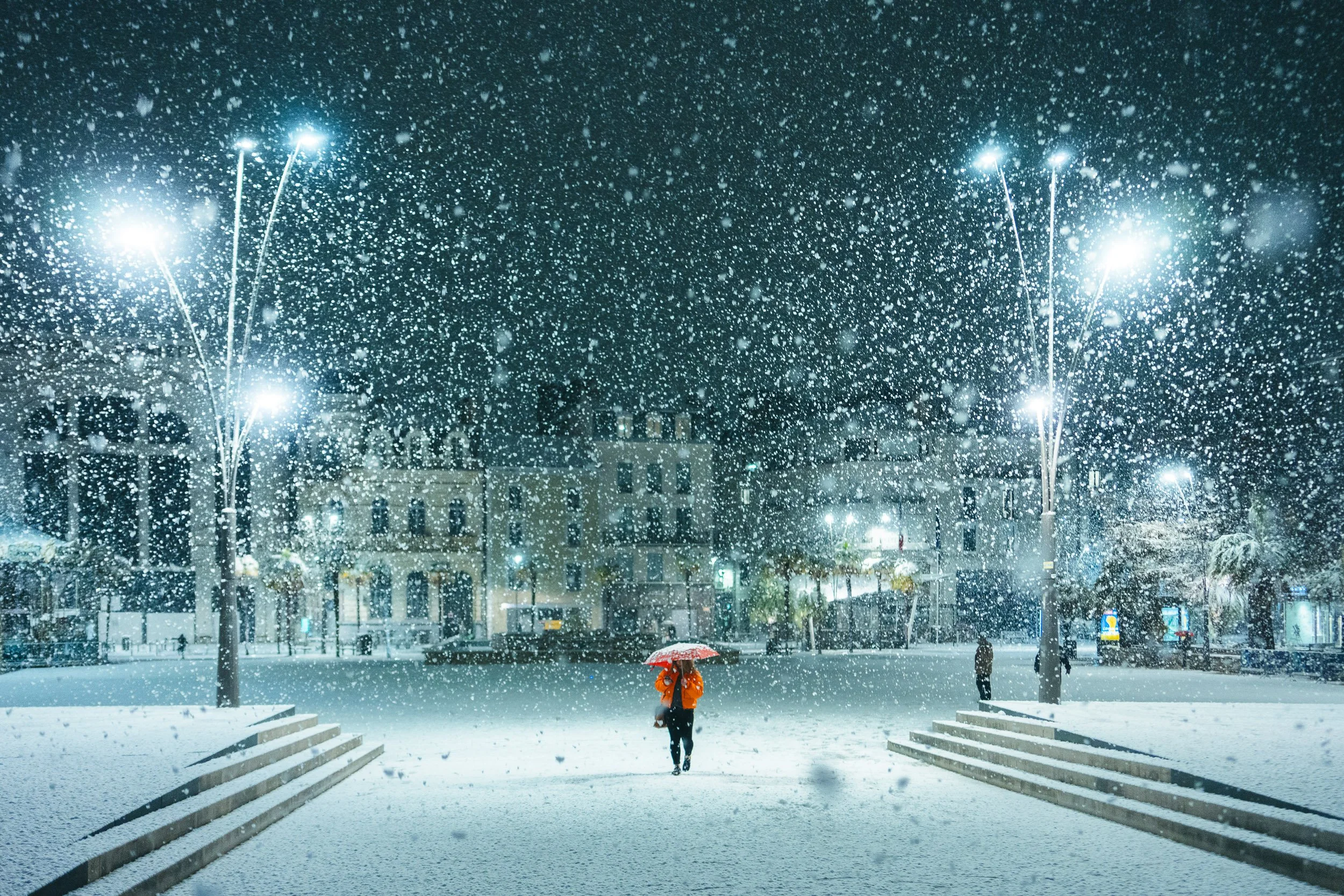 A person with an orange jacket and umbrella walking in a snowy city square at night, with snow falling heavily under bright streetlights and buildings in the background.