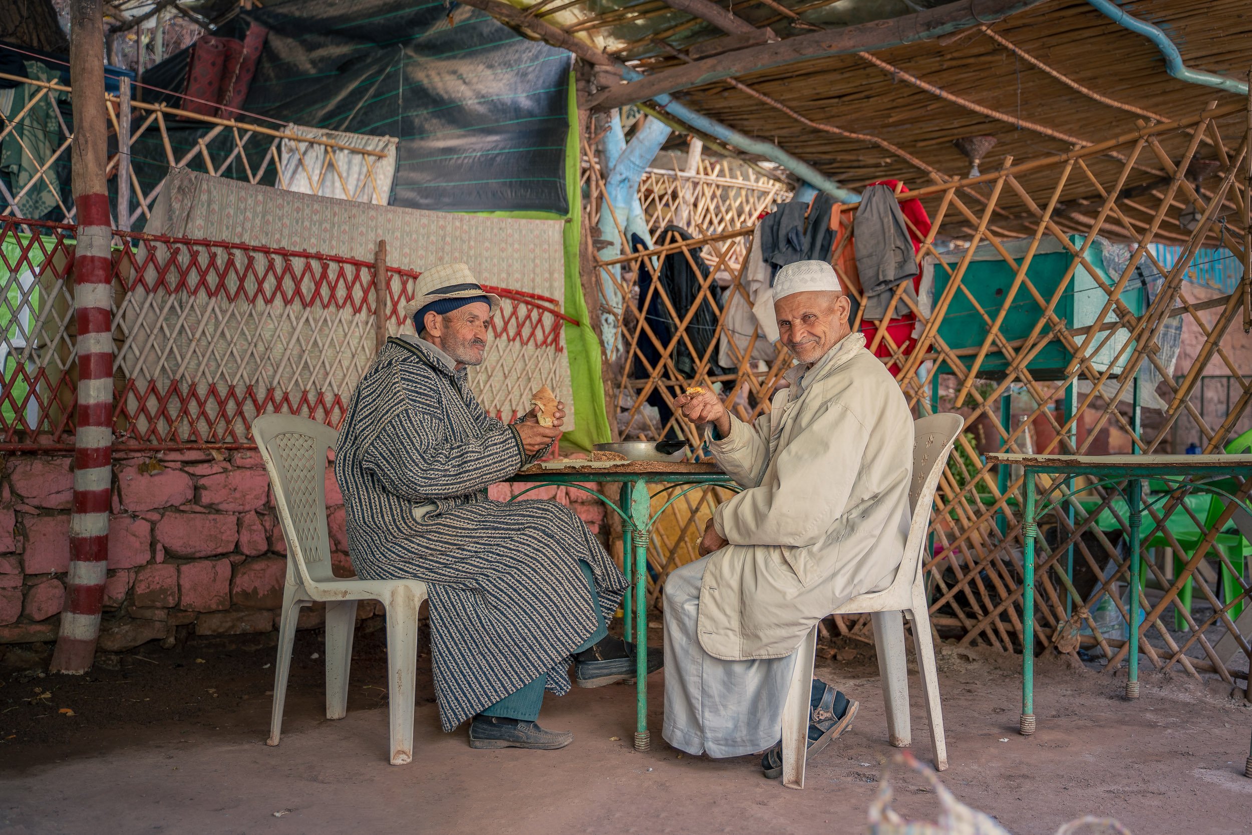 Two elderly men in traditional clothing enjoying a meal together outdoors, seated at a table with a simple metal top, under a makeshift shelter with woven bamboo walls and hanging laundry in the background.