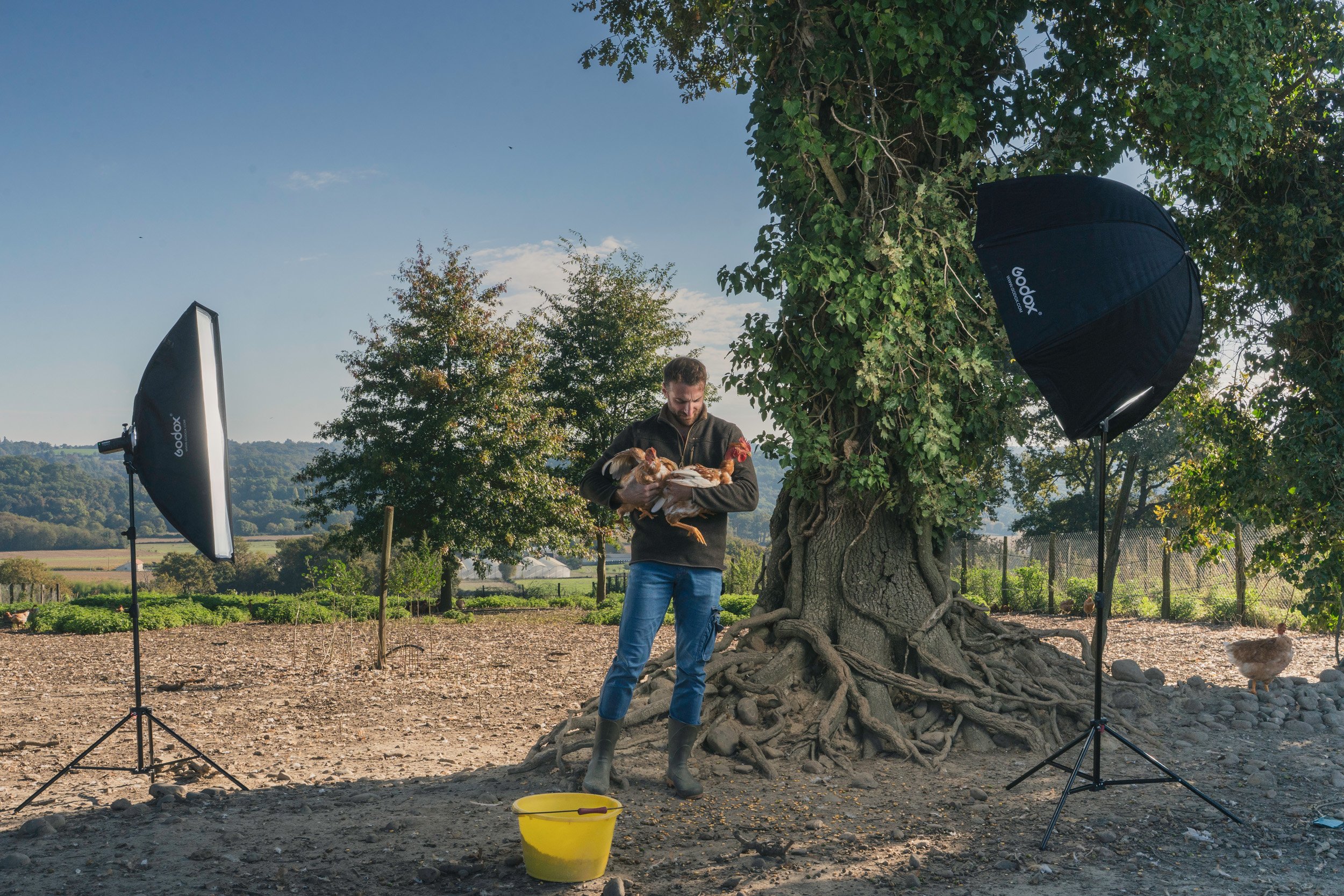 Man holding chickens in a farm setting with photography equipment and large trees.