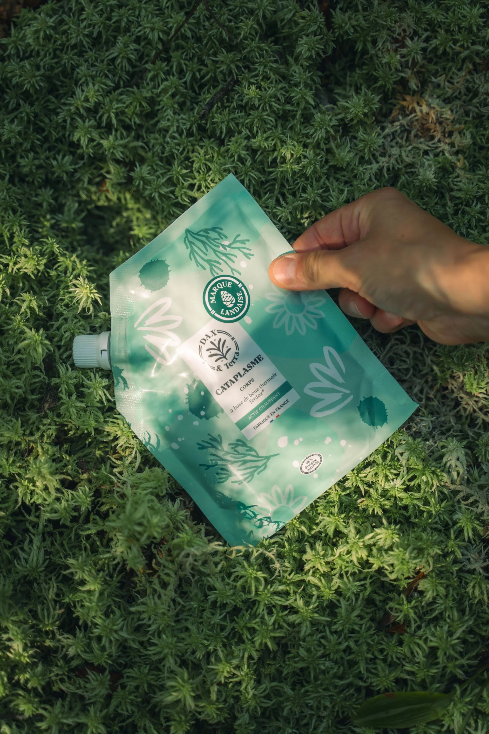A person's hand holding a green and blue refill pouch with a spout, placed on ground covered with small green plants, possibly in a garden or natural setting.