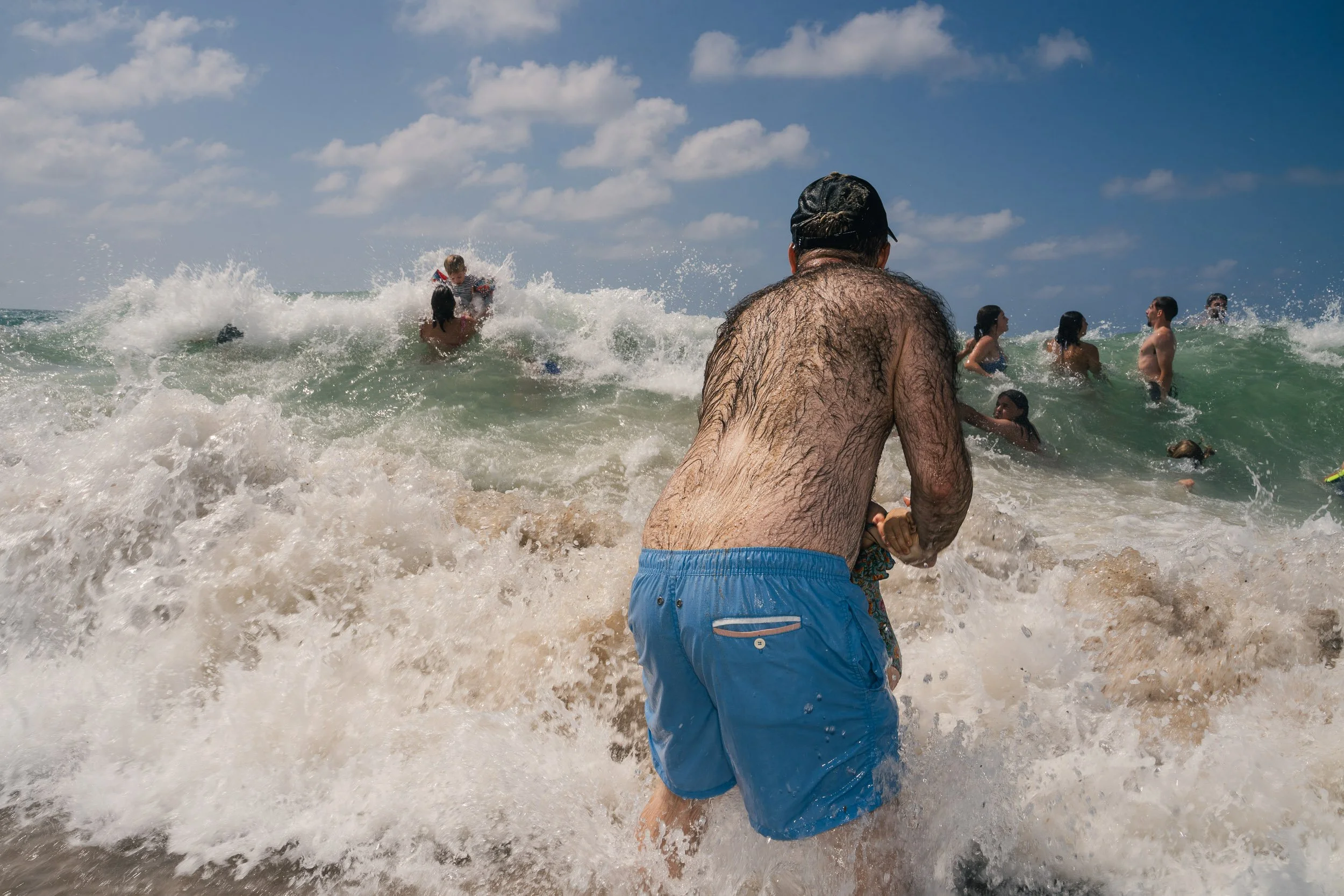 People swimming and playing in the ocean waves on a sunny day, with a man in blue swim trunks in the foreground facing away.
