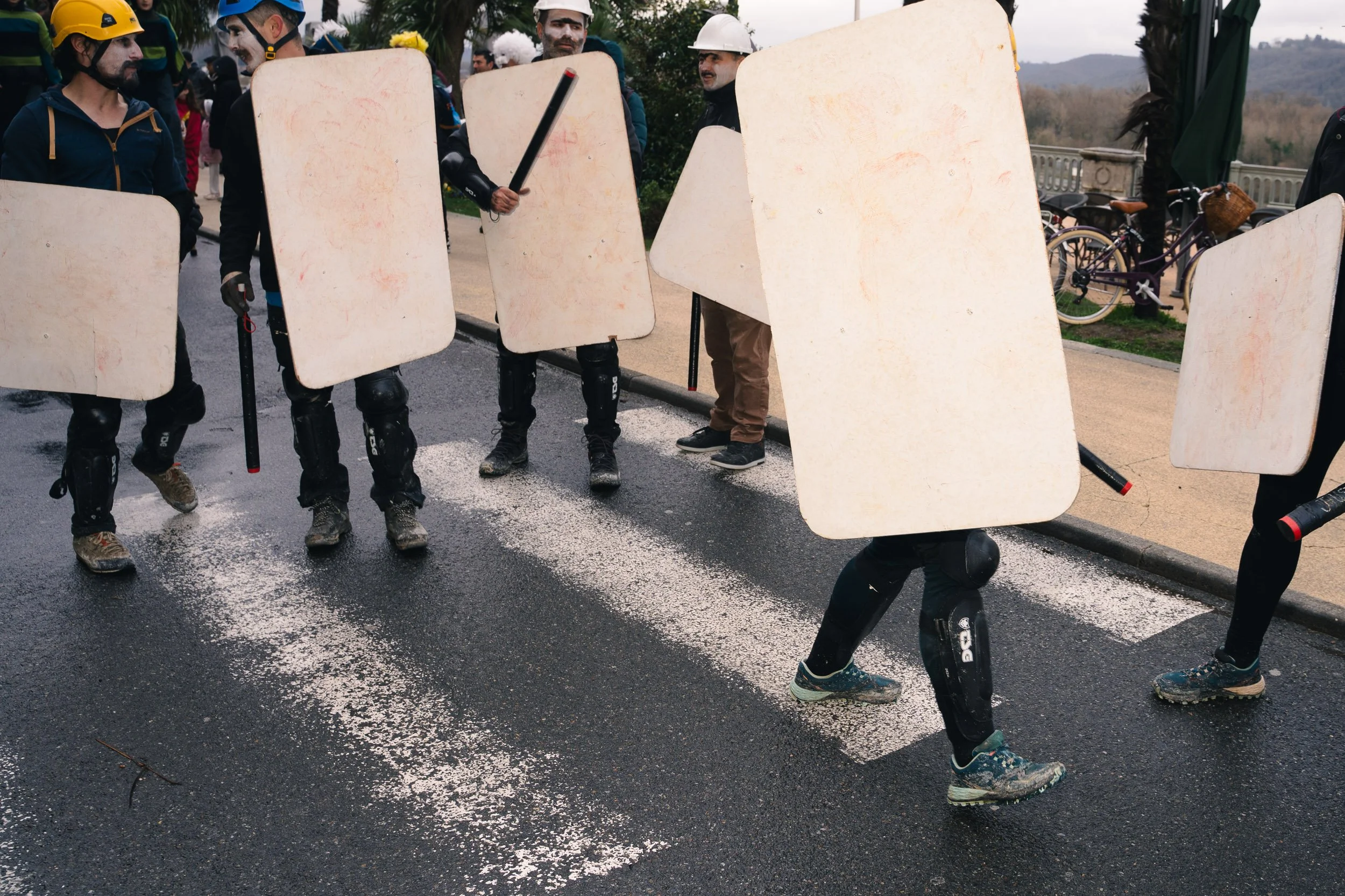 People dressed as riot police with shields and batons standing on a wet street during a protest or demonstration.