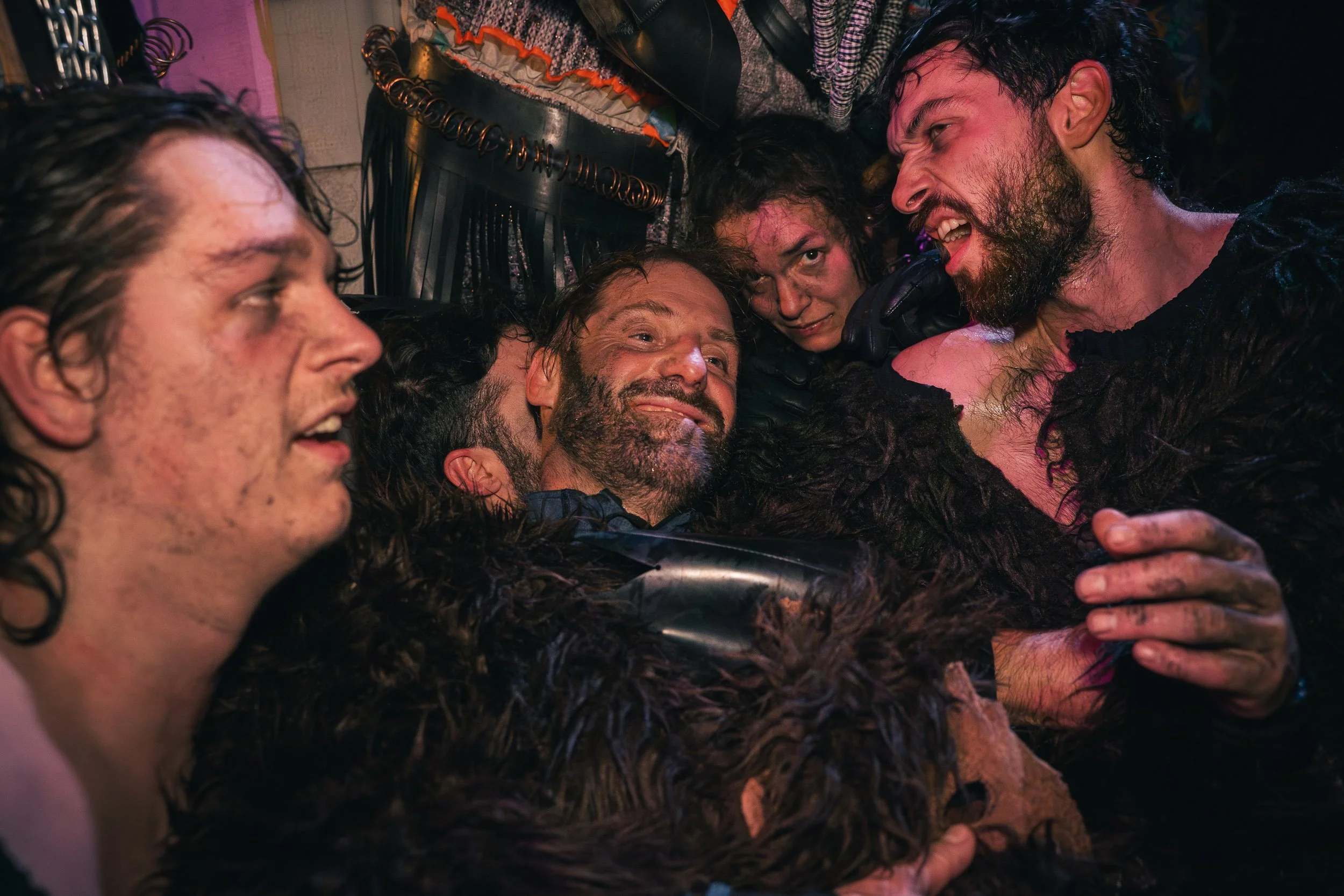 Group of five people in a close embrace, smiling and laughing, with some wearing black fur-like costumes, in a colorful setting.