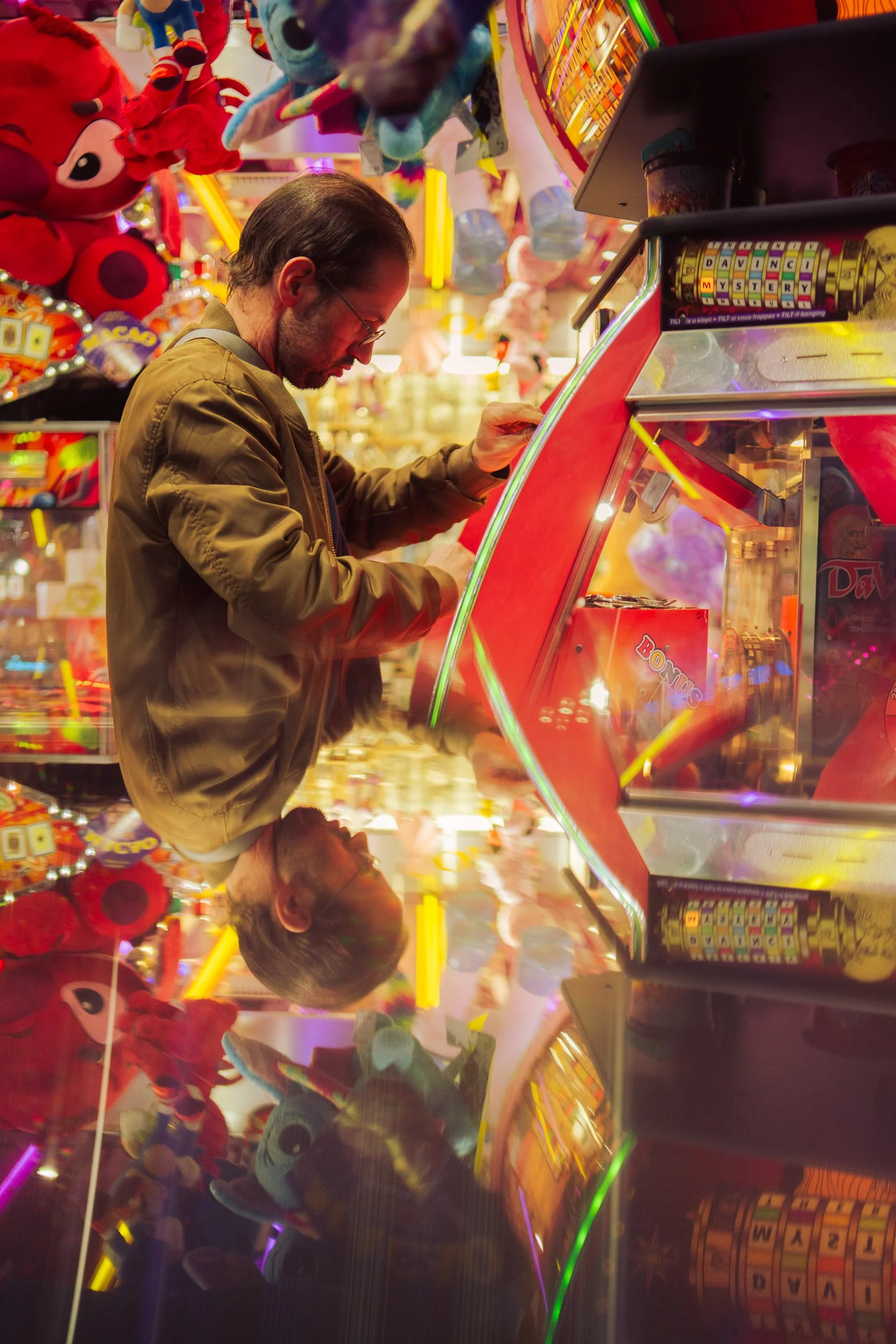 A man with glasses in a brown jacket playing a pinball machine in a colorful arcade filled with plush toys and neon lights. Reflections of the man and arcade are seen on the shiny surface below.