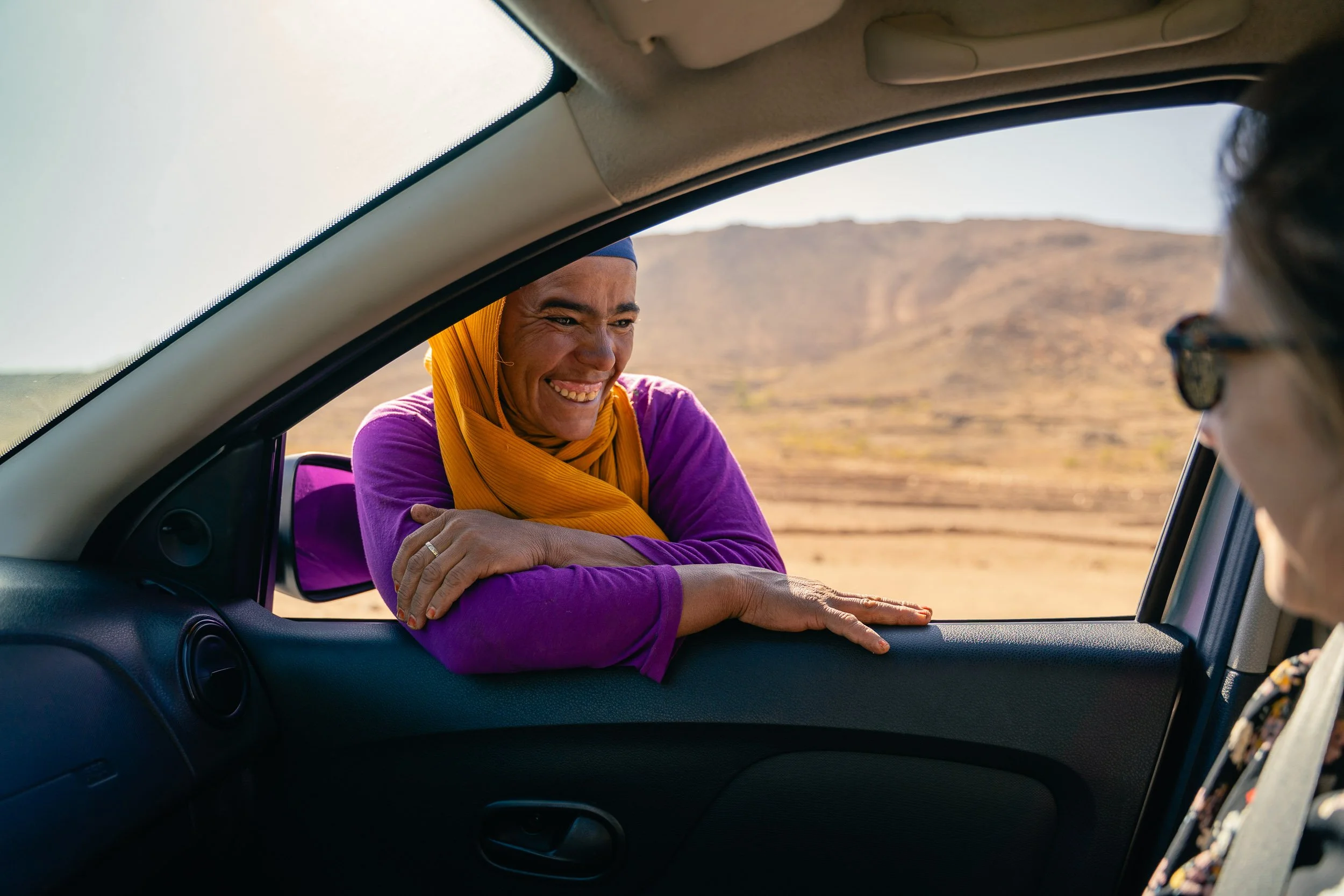 A woman in a purple shirt and yellow headscarf smiling and leaning on the car window, talking to a person inside the car with sunglasses, in a desert landscape.