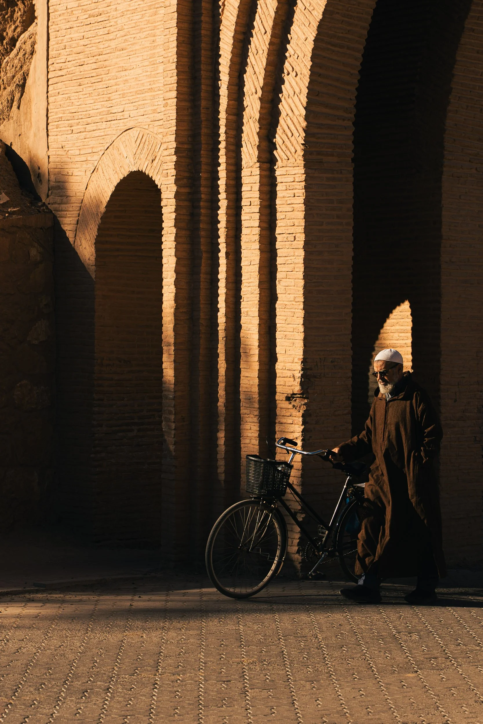 An elderly man with a white beard wearing dark sunglasses, a white cap, and a brown coat standing next to a black bicycle with a front basket, against a background of large brick arches in sunlight.