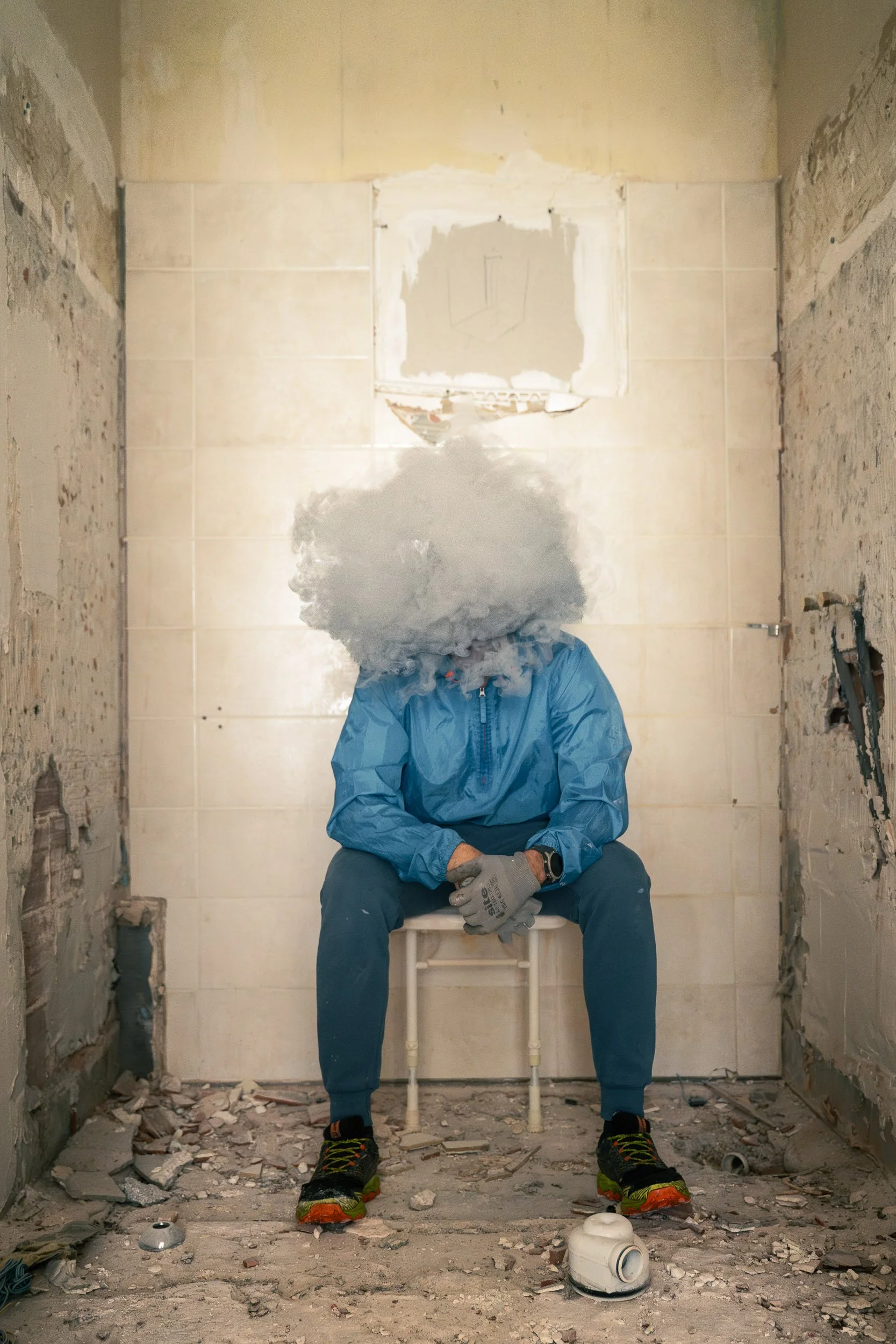 A person sitting on a small white stool with a cloud of smoke or vapor obscuring their face in a partially demolished room, wearing a blue jacket, gray gloves, and colorful sneakers, with debris on the floor.