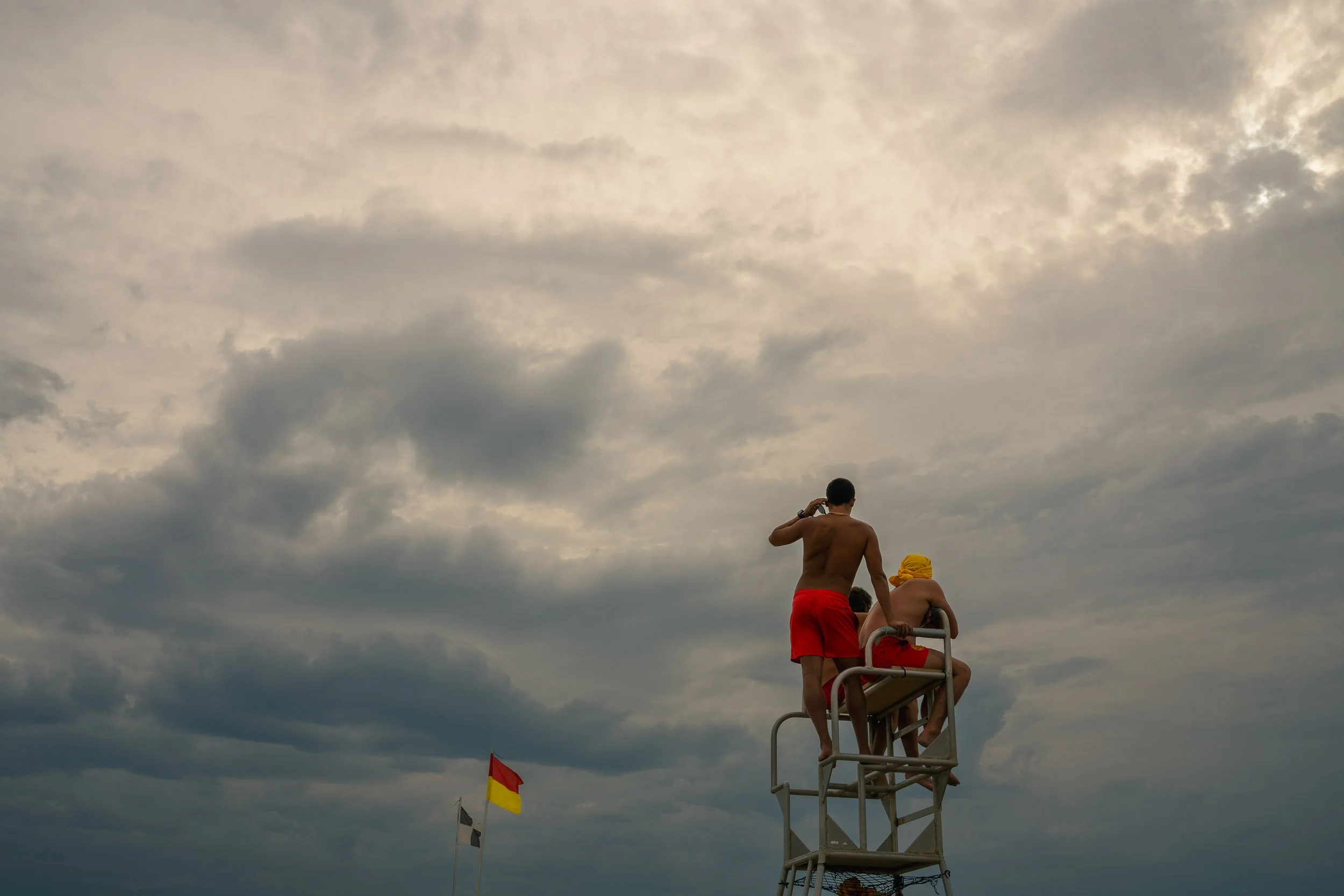 Three people sitting on a lifeguard stand at the beach, one talking on a phone and two sitting, with cloudy sky in the background.