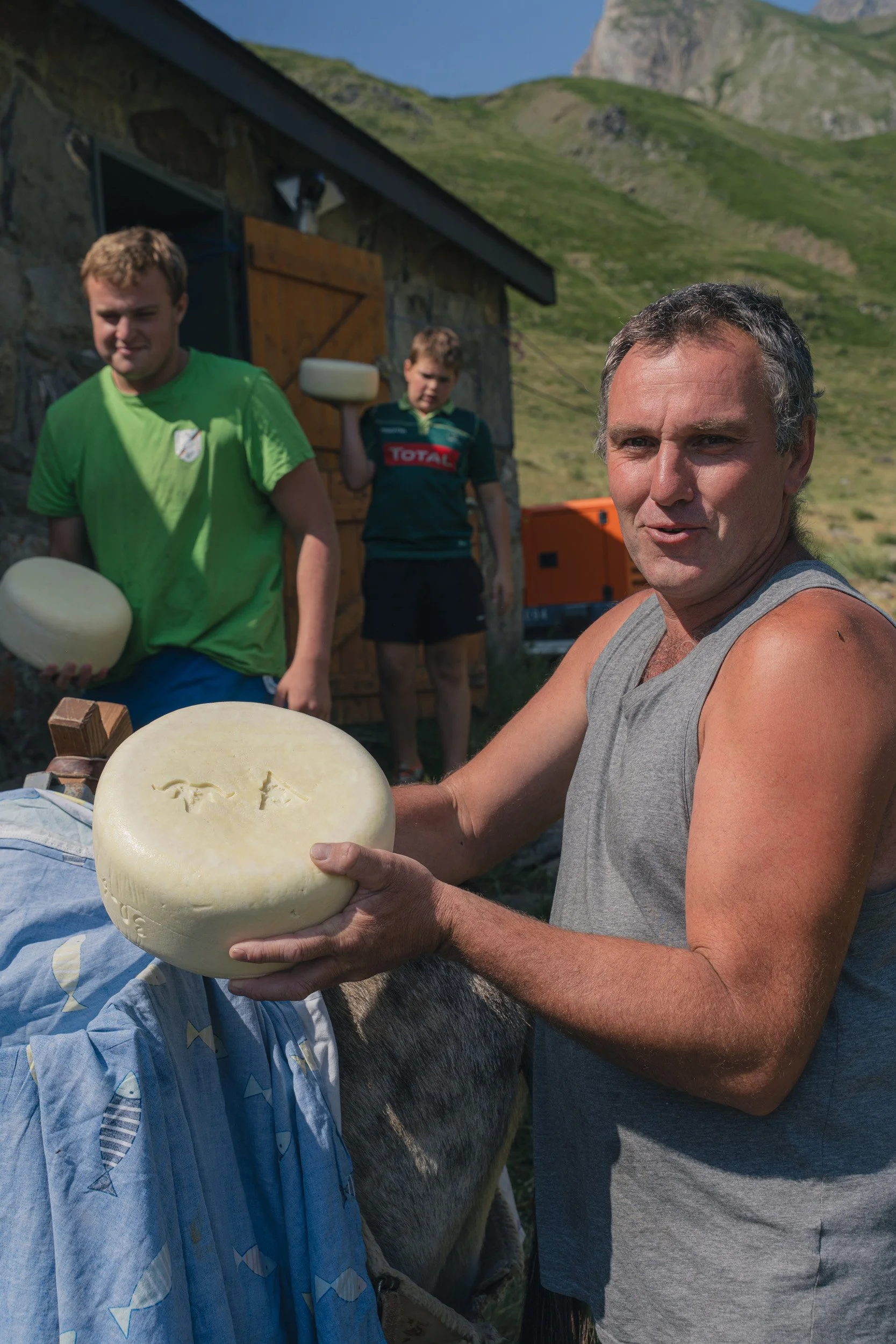 A man holding a large round cheese wheel with three cut marks, surrounded by a cow and two boys outdoors in a mountainous area.