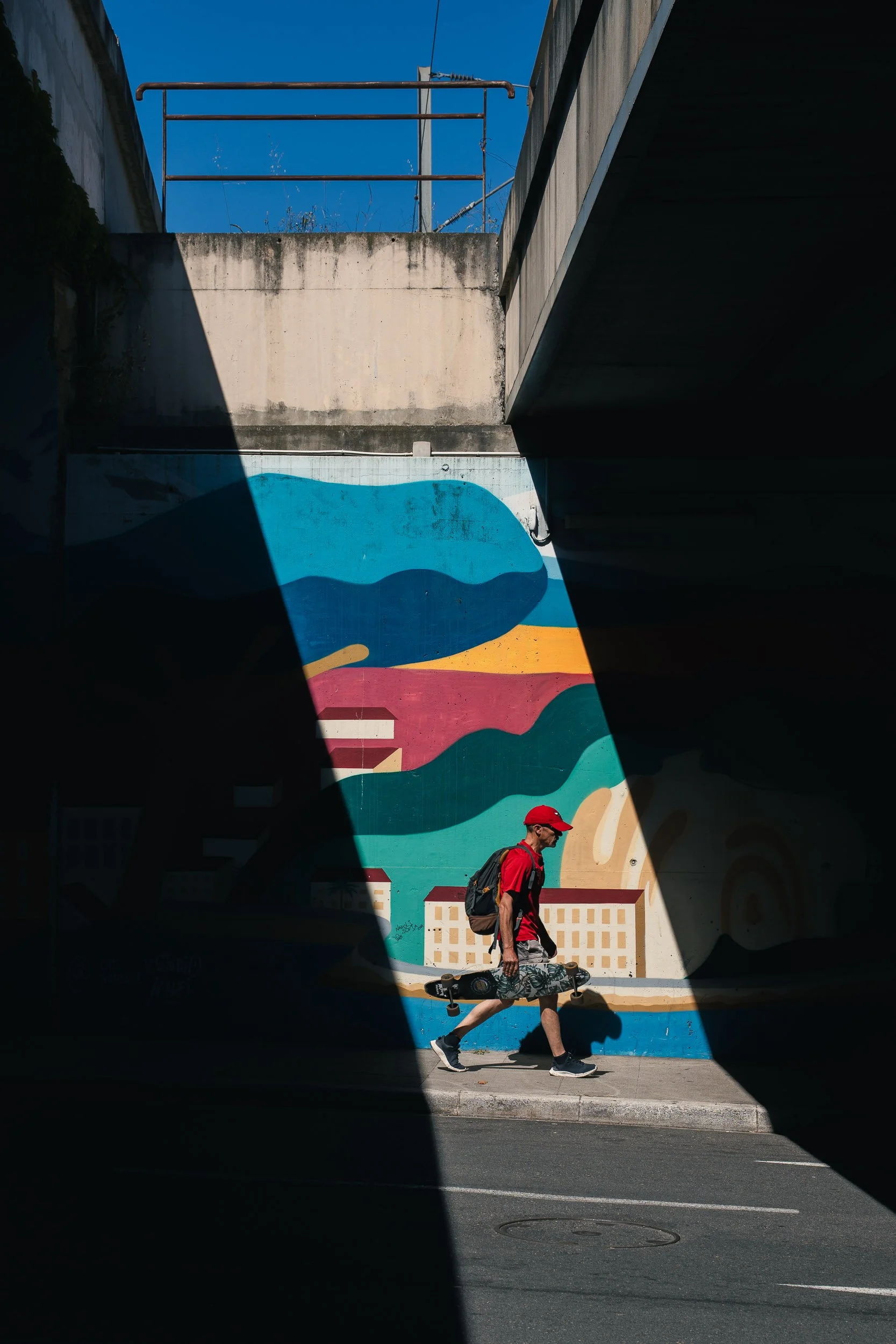 A man walking with a skateboard under a bridge with a colorful mural of mountains and buildings painted on the wall, wearing a red cap, red shirt, black shorts, and carrying a backpack.